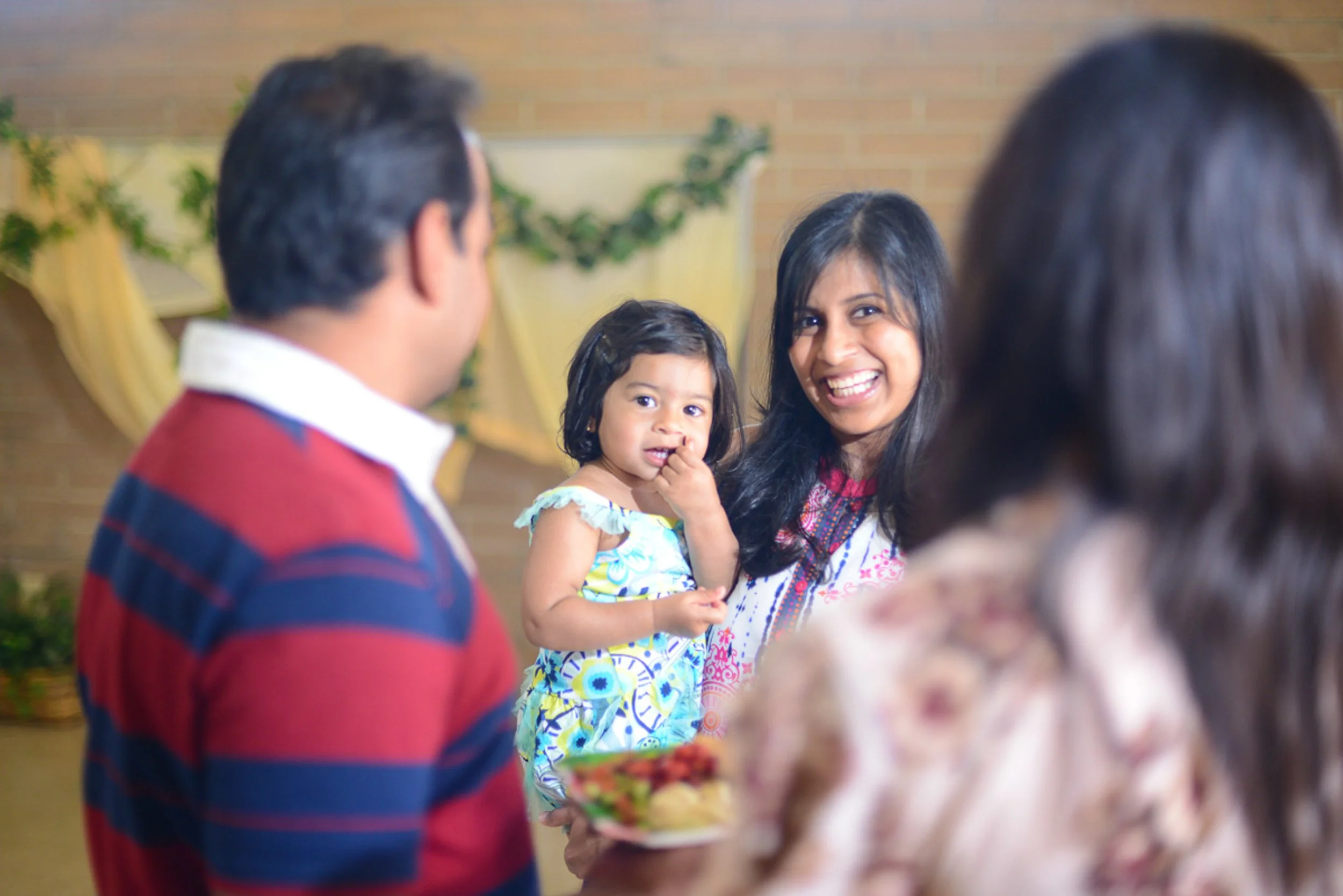 Group of people at a family gathering, including a woman holding a young girl, smiling. Others are partially visible in the foreground. The background has decorations and plants.