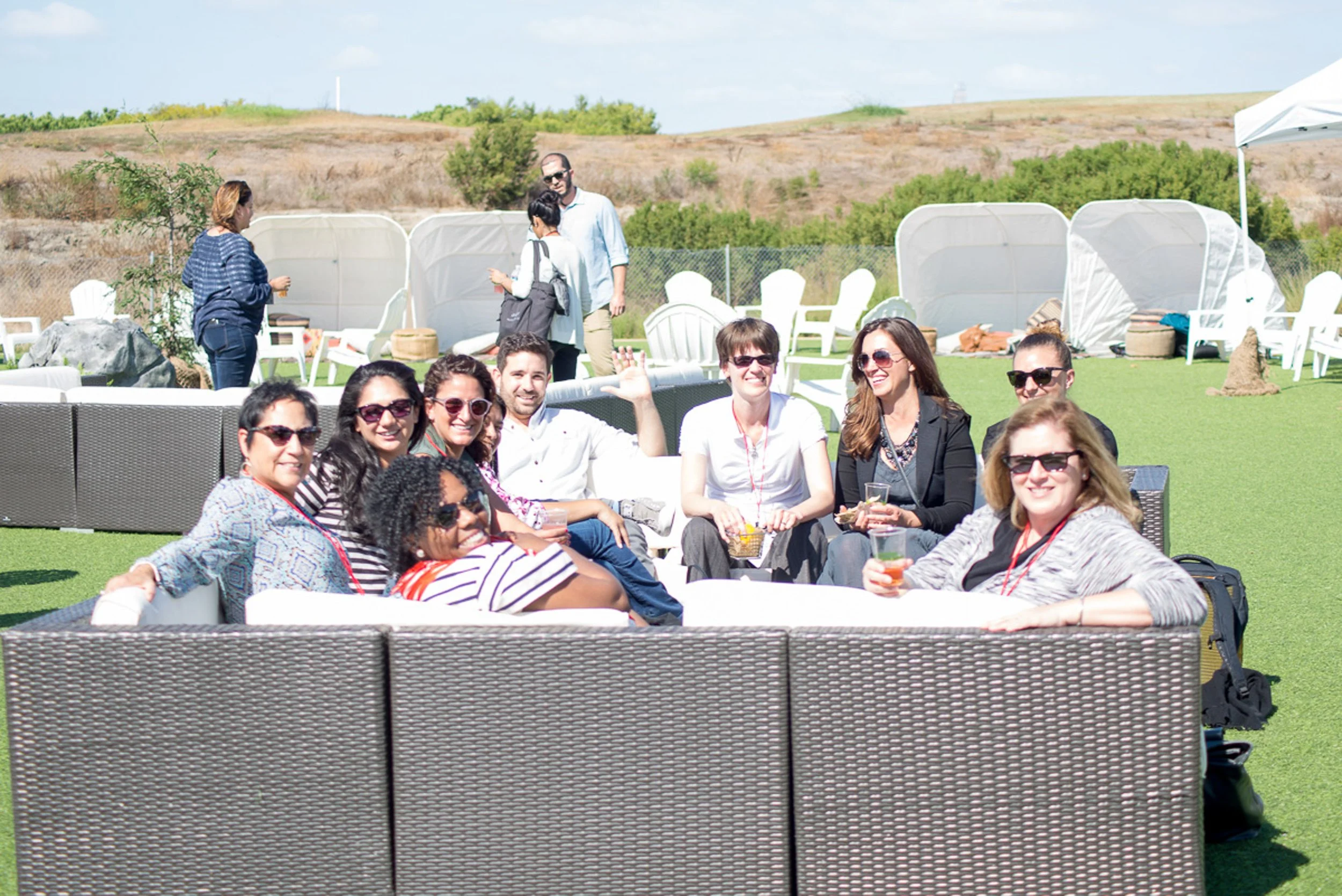 A group of people sitting on outdoor wicker furniture at a social gathering, smiling and holding drinks, in a sunny outdoor setting with hills and other people in the background.