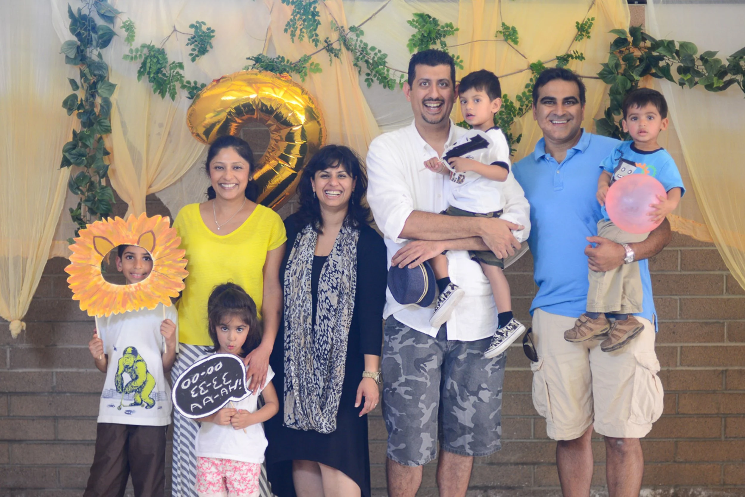 Event photography. Group of seven people, including adults and children, celebrating with balloons and props in front of a decorated backdrop with greenery and hanging fabric
