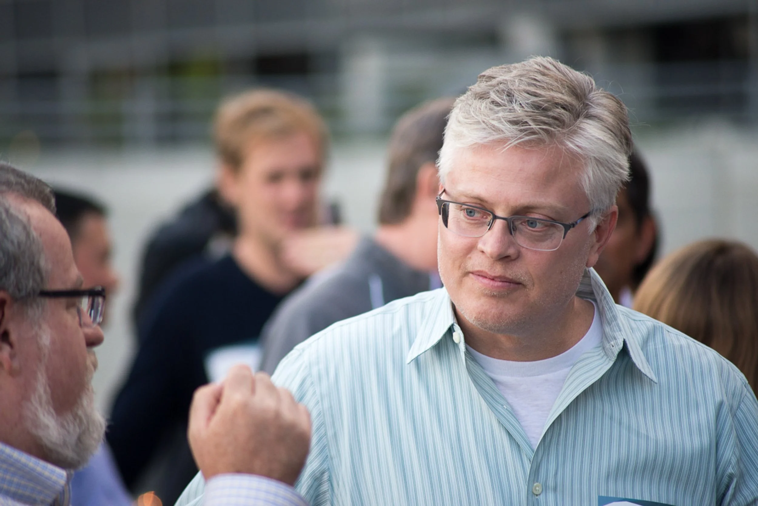 Event photography. A man with glasses and graying hair in a light blue striped shirt is outdoors, looking to the side, surrounded by other people in conversation.