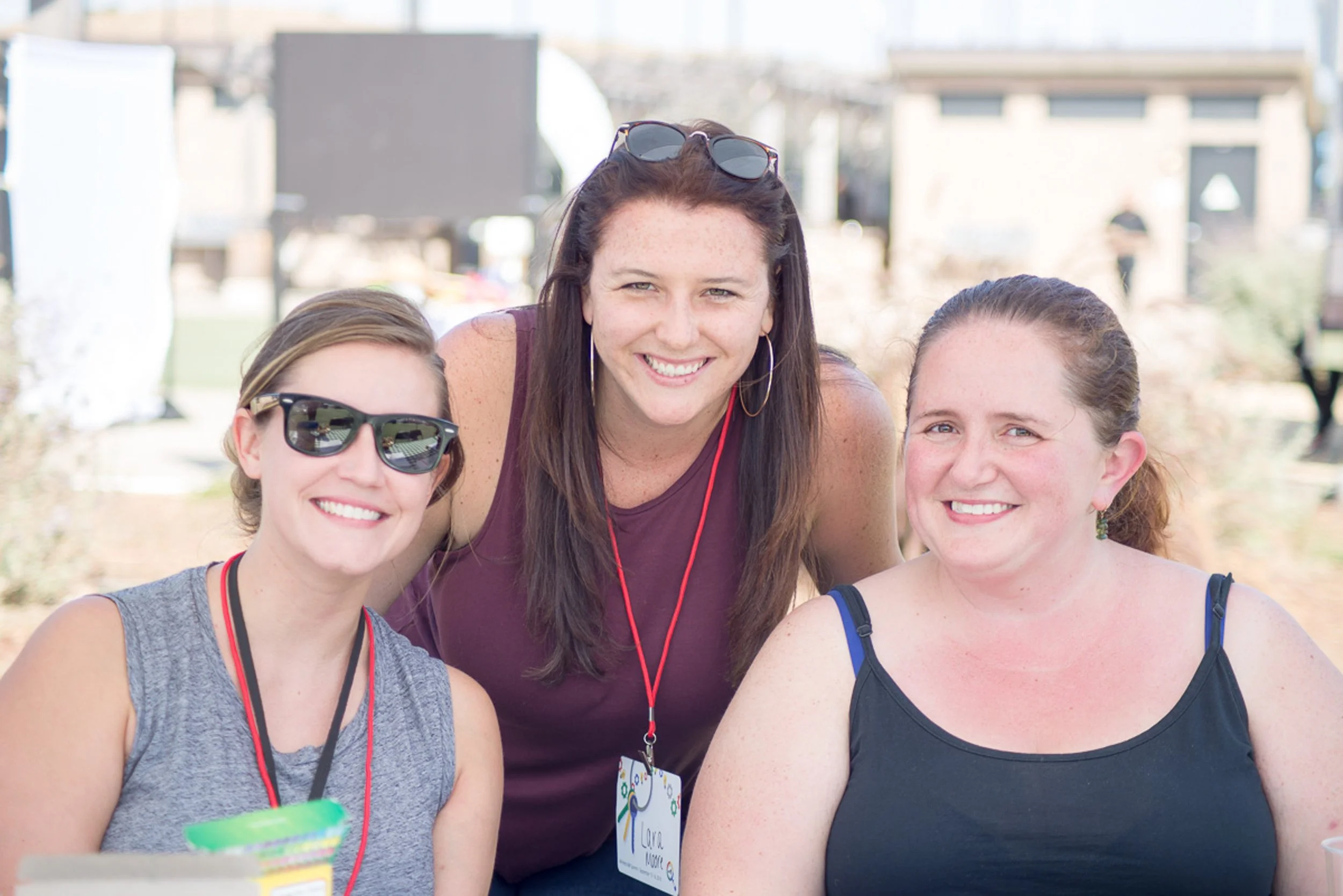 Three women smiling at an outdoor event. The woman in the center has long dark hair, sunglasses on her head, and a purple top. The woman on the left has short hair, wearing sunglasses and a gray sleeveless top. The woman on the right has brown hair p