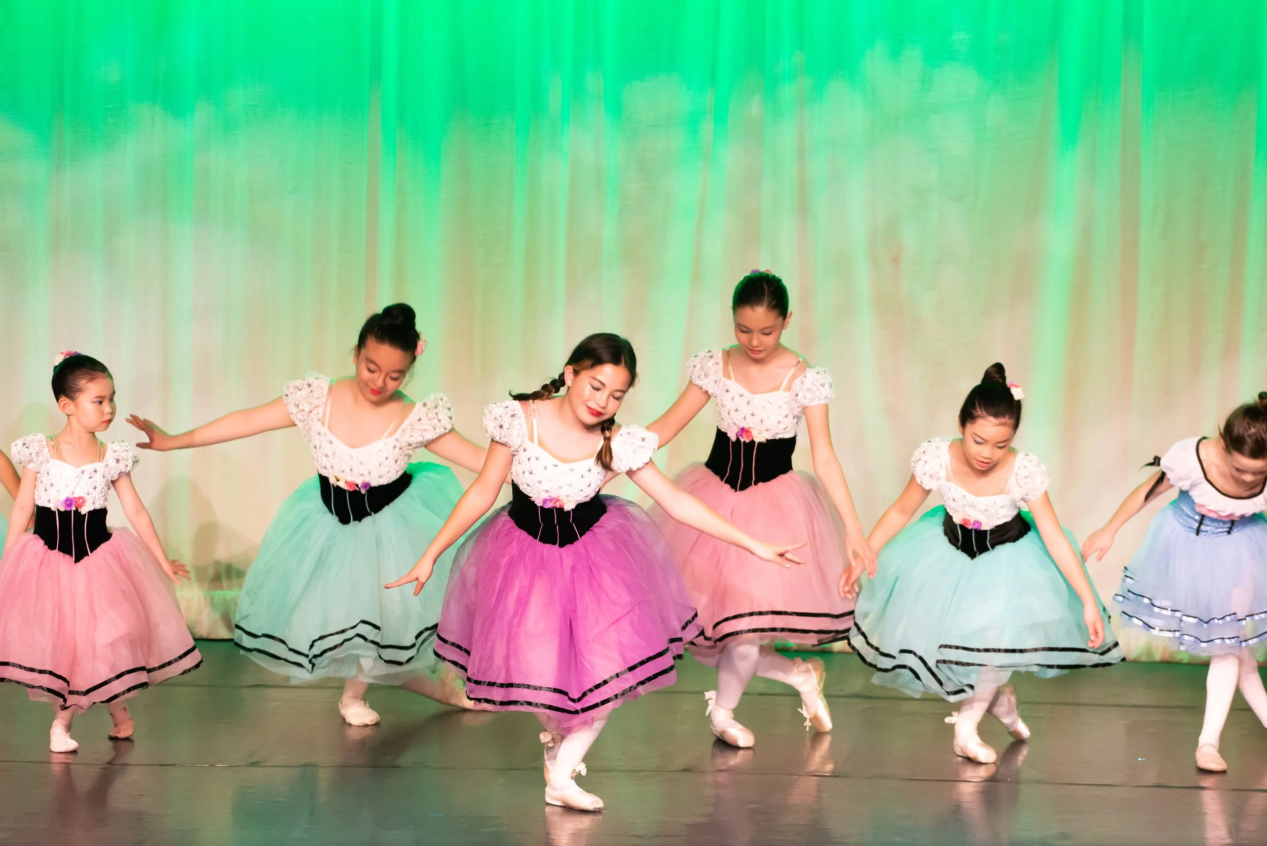 School photography. Young girls performing a ballet dance on stage, wearing colorful tutus and white tops with floral accents, with a green illuminated background.