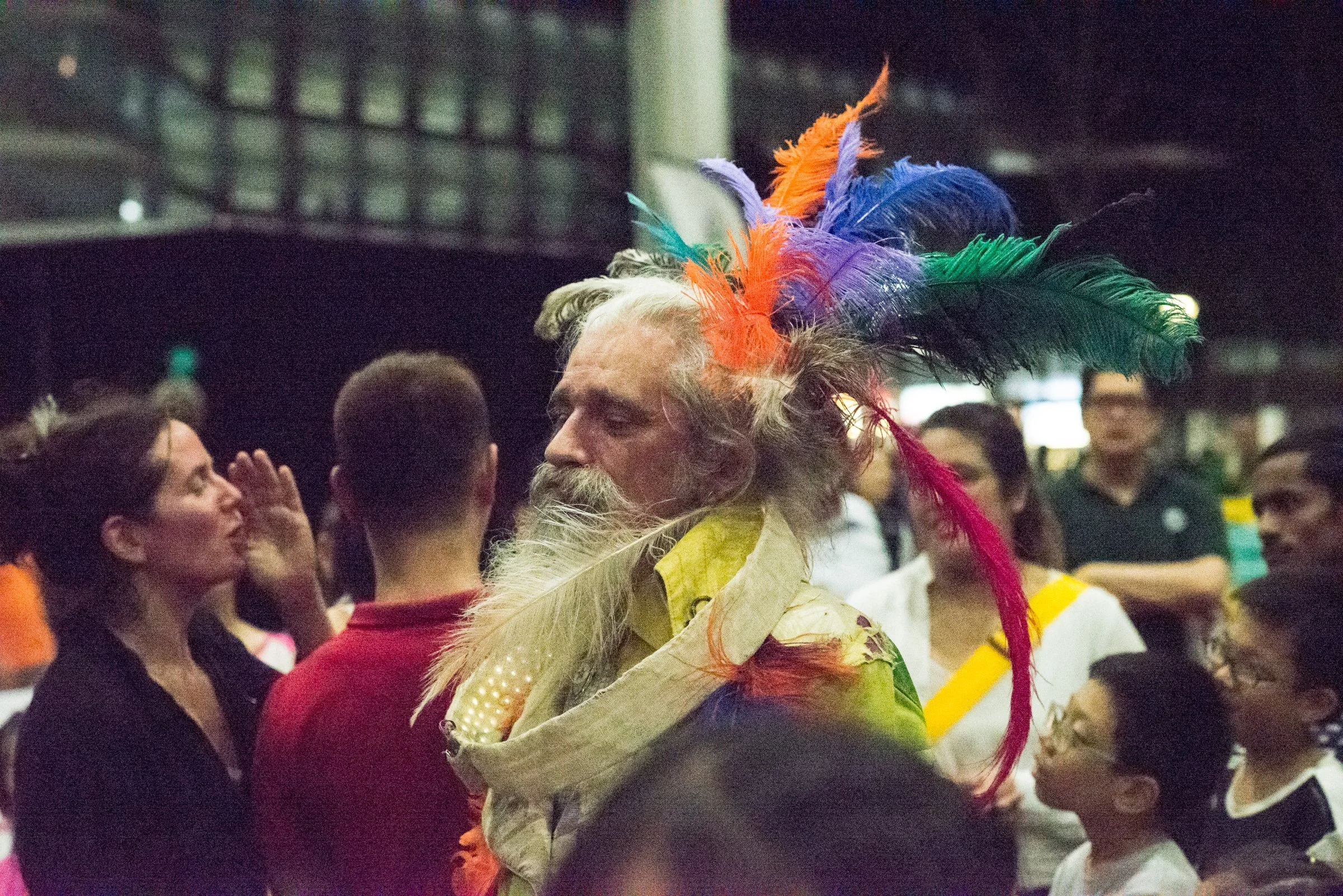 A man with long gray hair and beard dressed in colorful, feathered costume at a crowded indoor event.