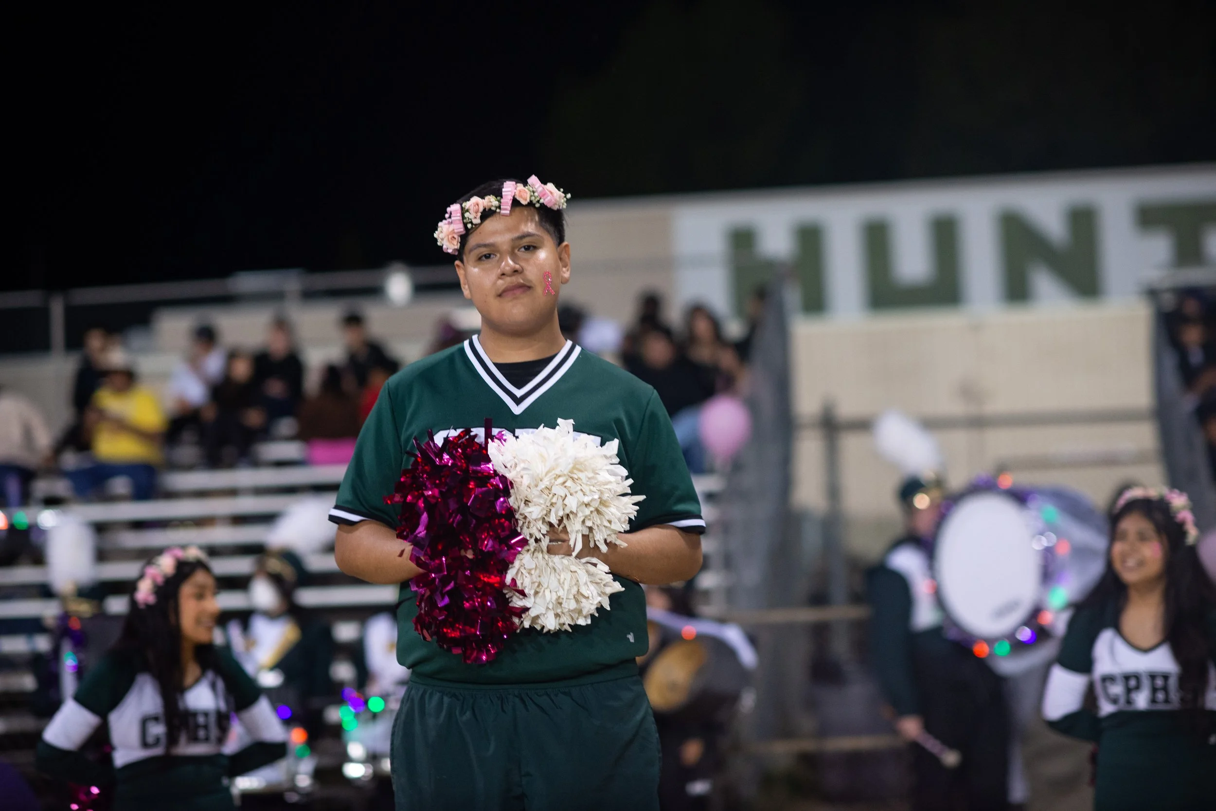 School photography. A young person dressed in a green sports jersey holding pom-poms, wearing a flower crown, at a nighttime school event with people sitting on bleachers and a marching band in the background.