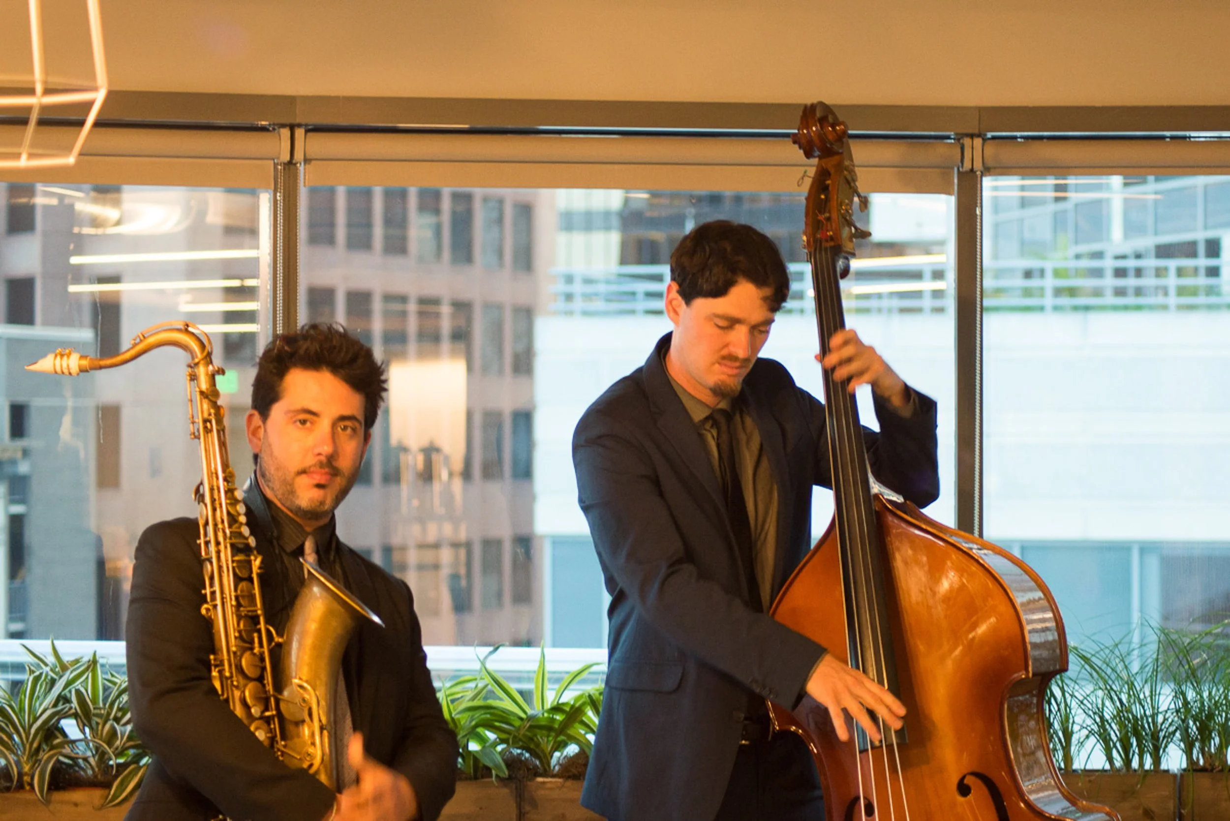 Event photography. Two musicians in suits playing jazz instruments: one with a saxophone, the other with a double bass, indoors with city buildings through windows in the background.
