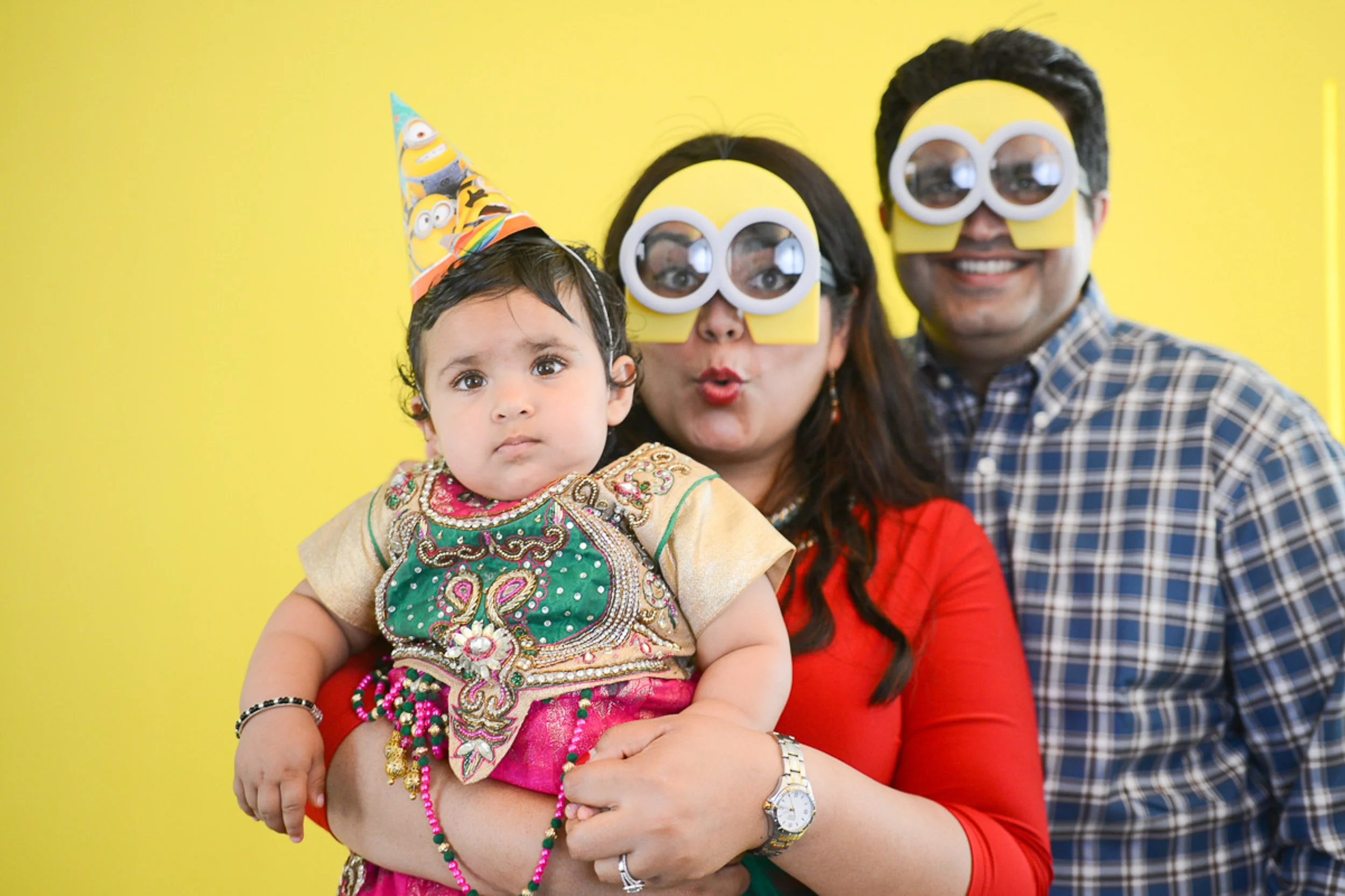 A family of three celebrating a birthday, with the mother holding a young girl, and both the mother and the man wearing yellow Minion-themed glasses. The girl is wearing a colorful party hat and traditional dress, while the background is yellow.