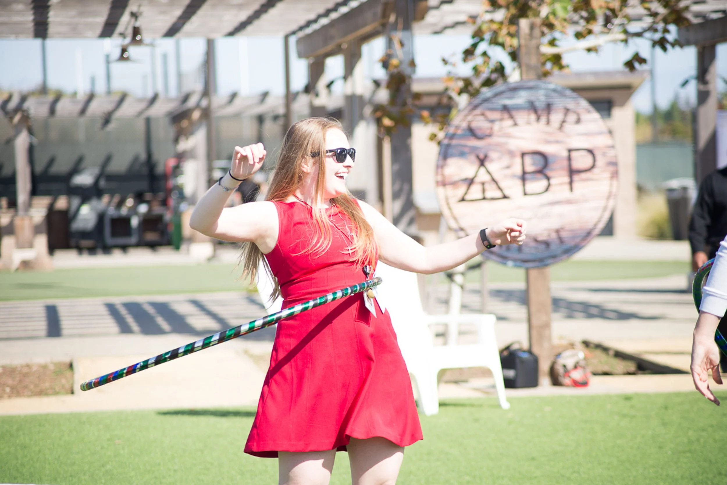 A woman in a red dress and sunglasses dancing outdoors at a college event, with a Greek lettered banner in the background.