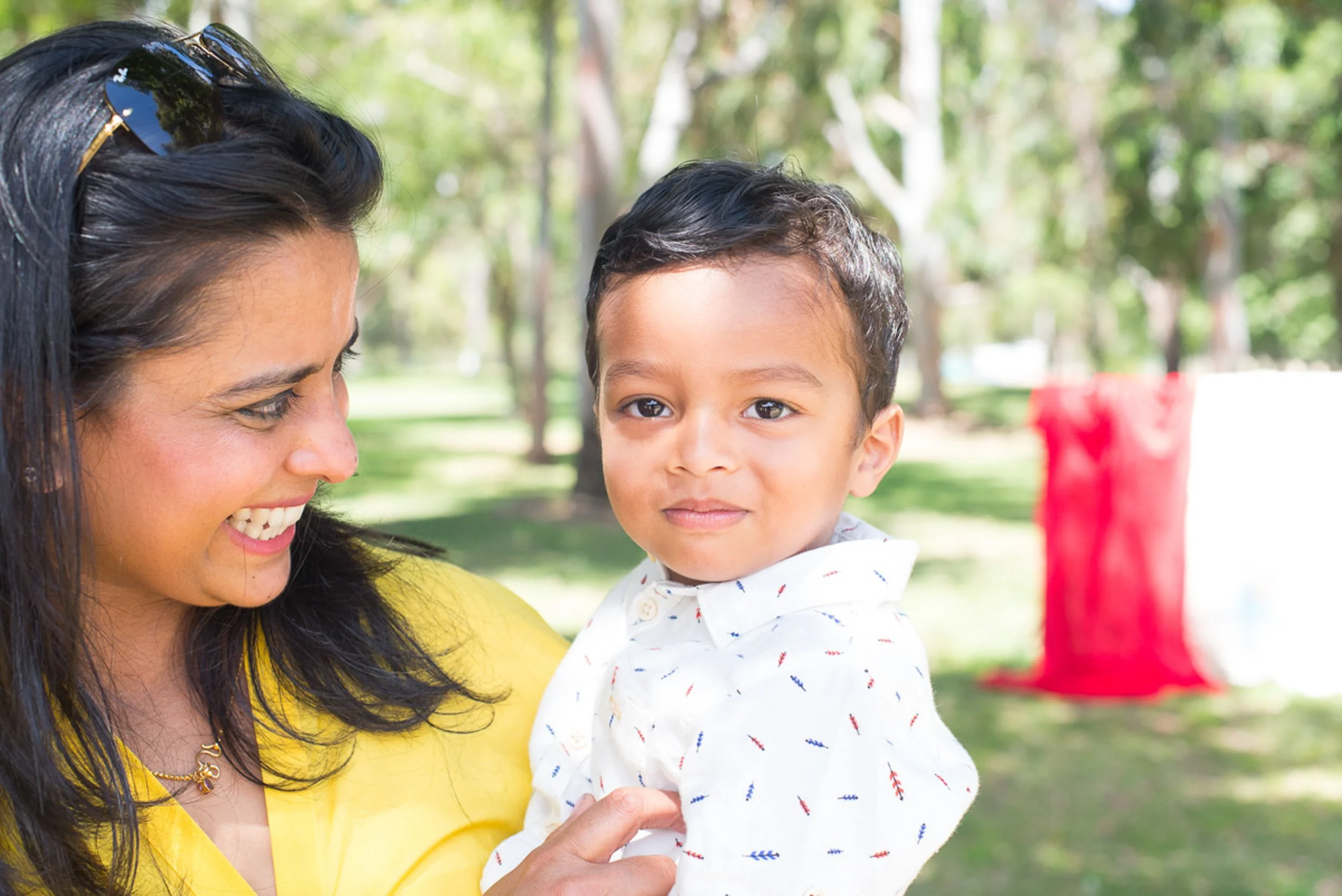 A woman smiling and looking at a young boy outdoors in a park with trees in the background.