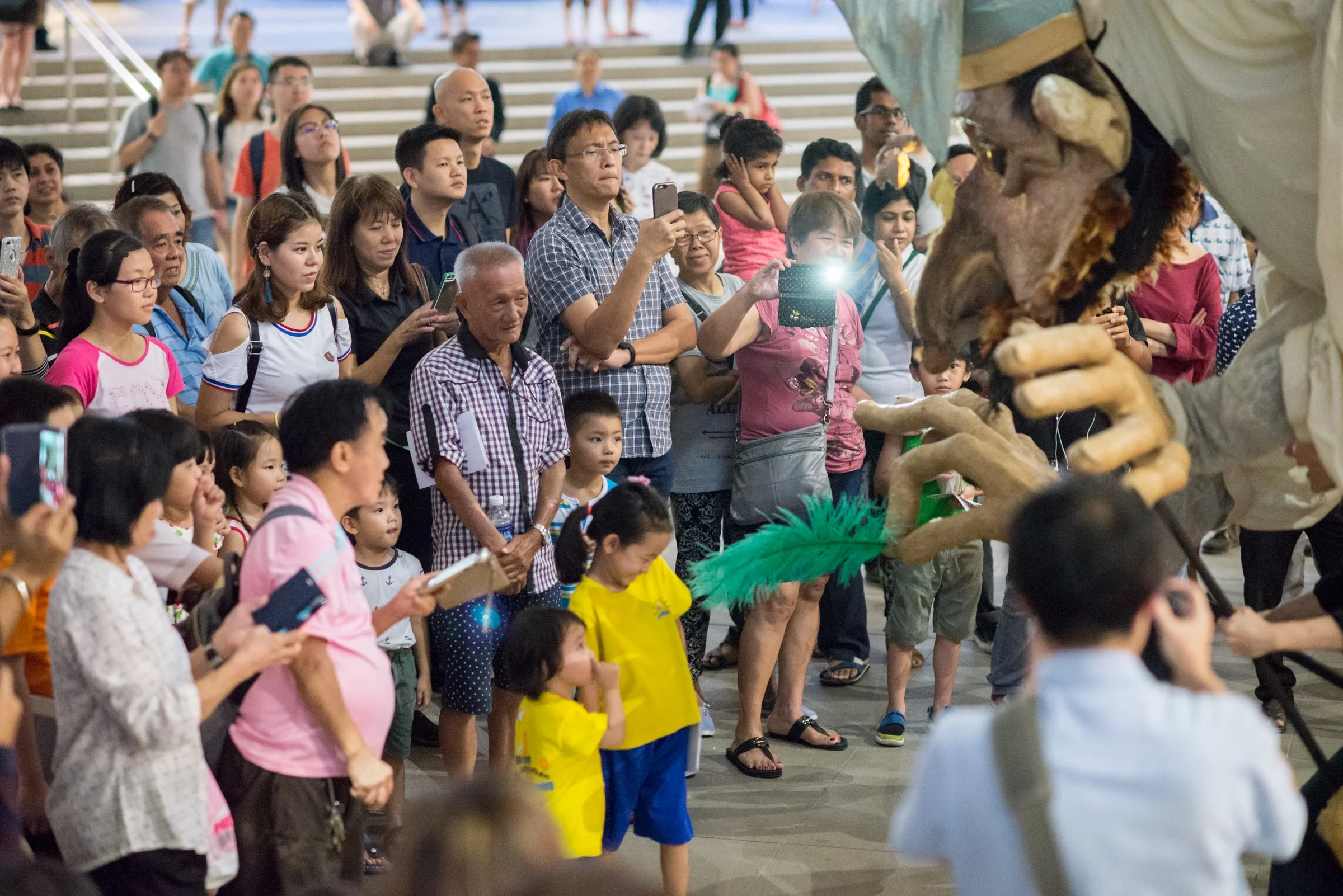 A group of children and adults watching a street performance with a person wearing a long nose mask and holding a large puppet or prop in front of a crowd.