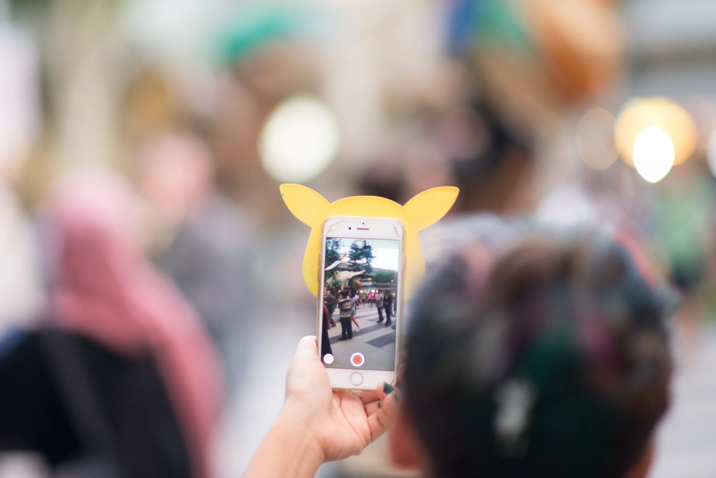 Person taking a photo with a phone on a yellow Pikachu-shaped phone holder at an outdoor event.