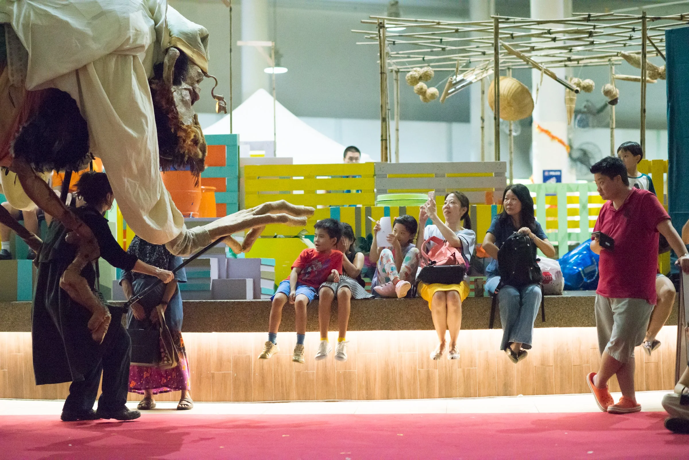 A puppet show featuring an elderly puppet with long, projecting nose and pointed ears, operated by puppeteers, with an audience of children and adults watching and taking photos in a colorful indoor venue.