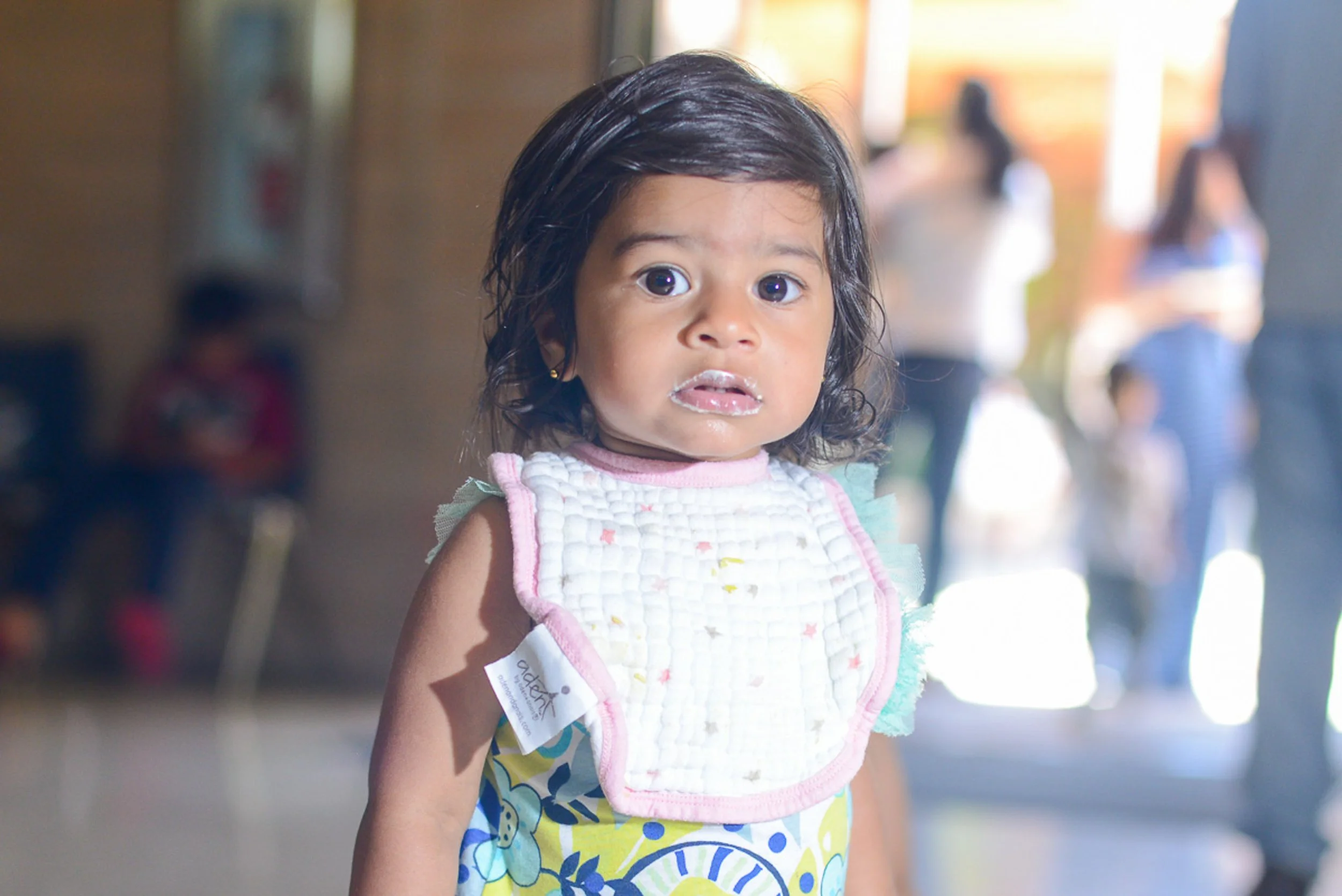 Event photography. Young girl with short dark hair and wide eyes, wearing a colorful dress and a white bib with pink trim, standing indoors with blurred figures in the background.