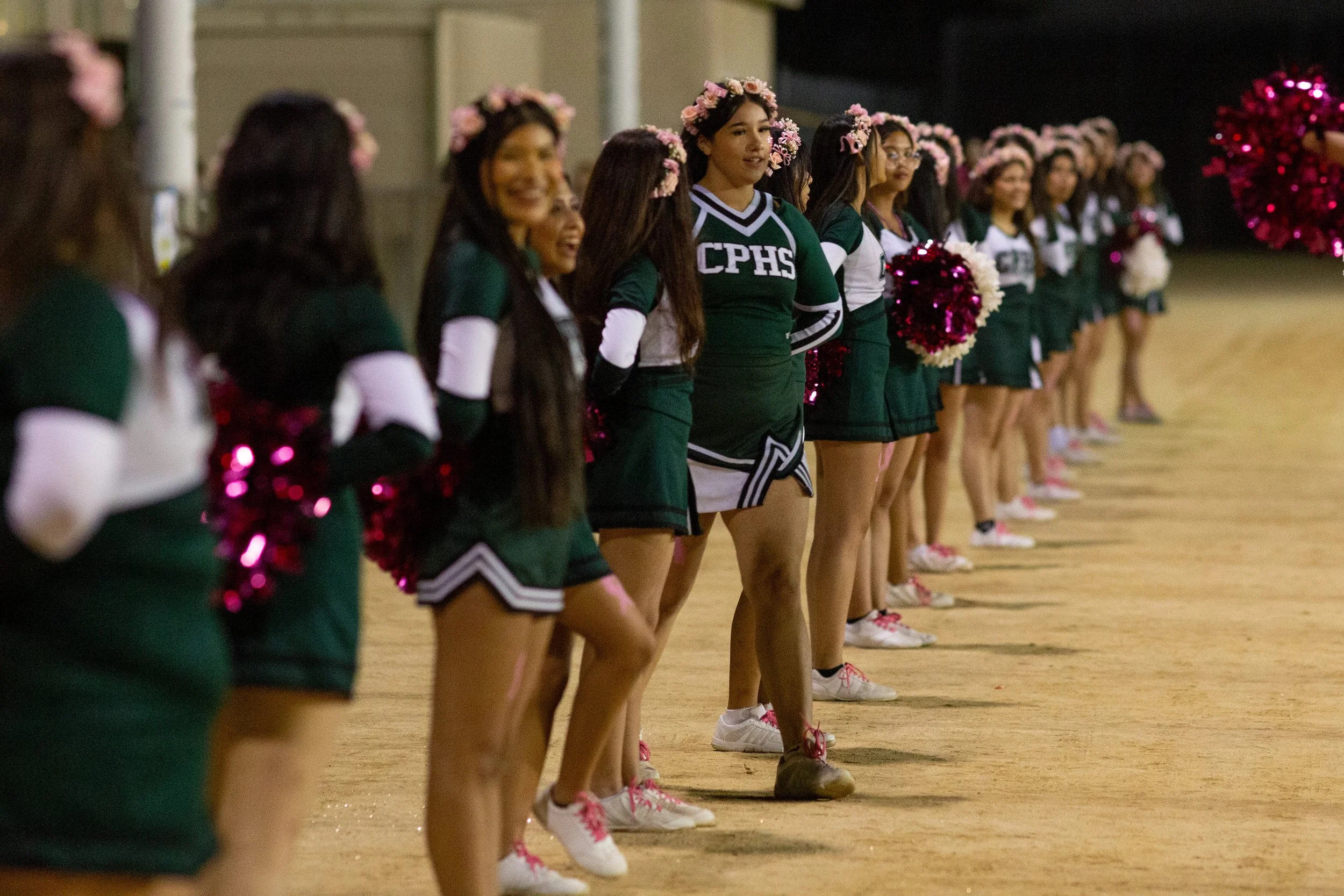 School photography. Line of cheerleaders in green and white uniforms standing on a field at night, holding pink and white pom-poms, with some wearing flower crowns.