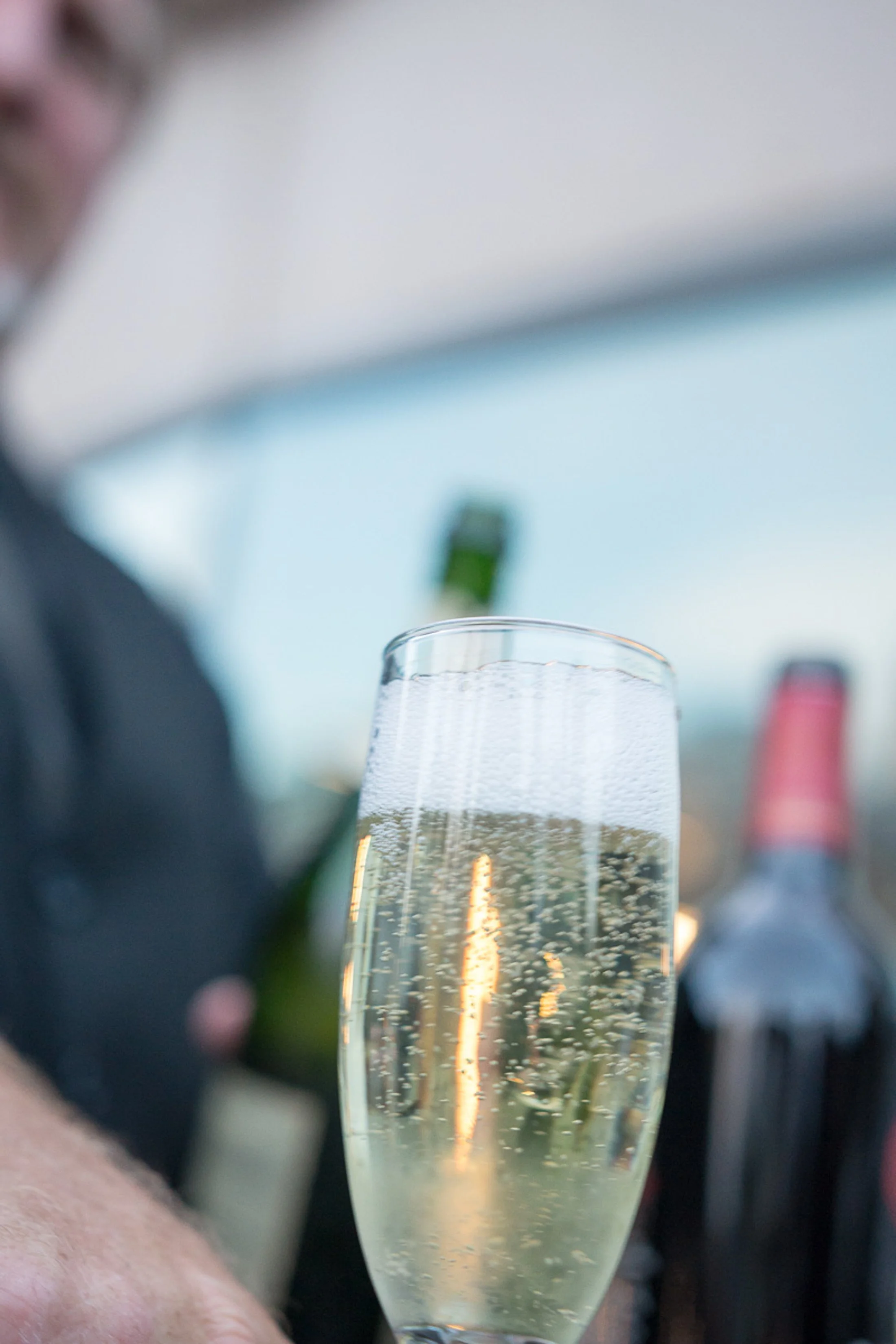 Event photography. A close-up of a glass of sparkling champagne with bubbles, with blurred bottles and a person in the background.