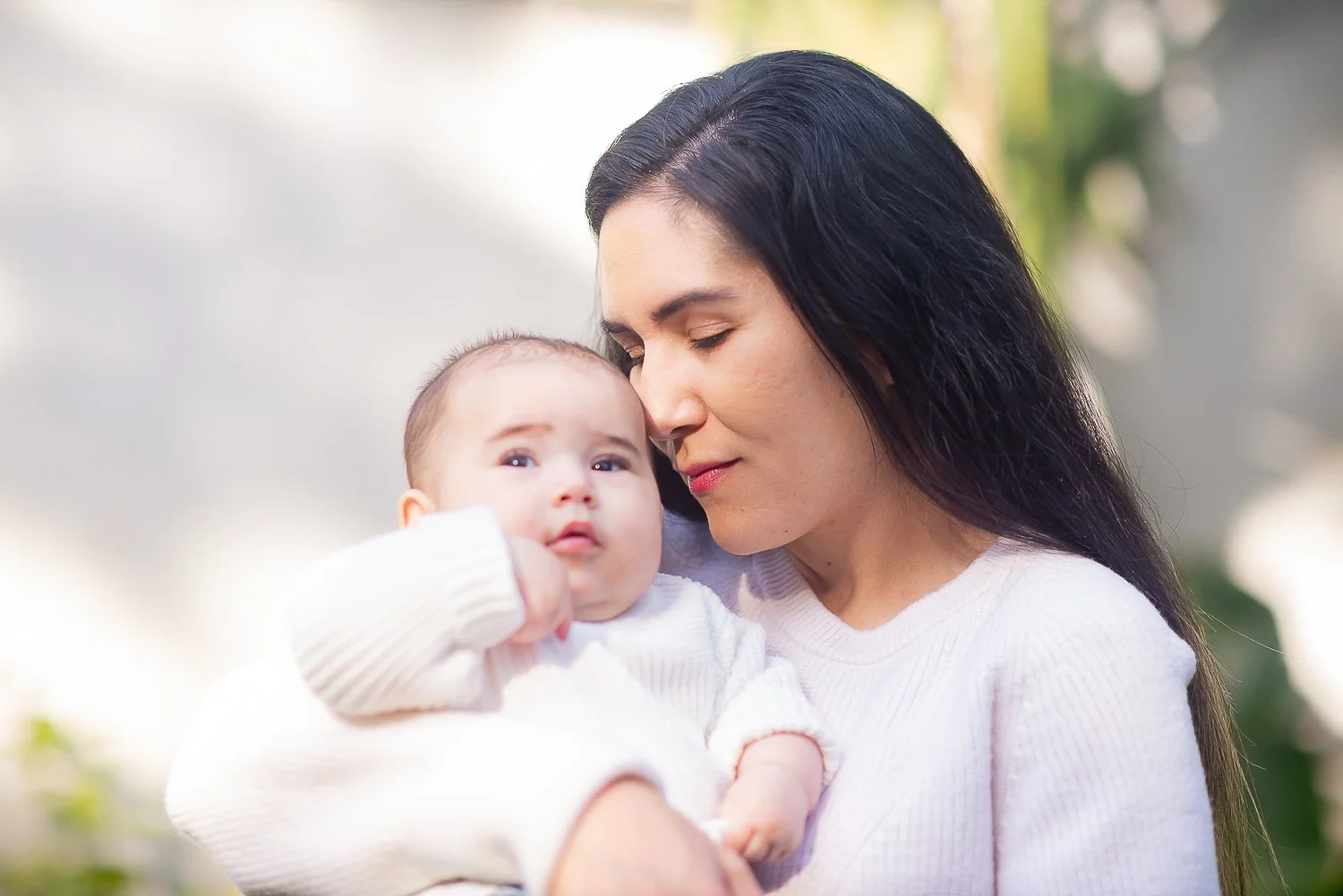 A woman holding a baby outdoors with a blurred background, the woman with long dark hair and closed eyes, the baby with light hair and wide eyes, both dressed in light-colored clothing.