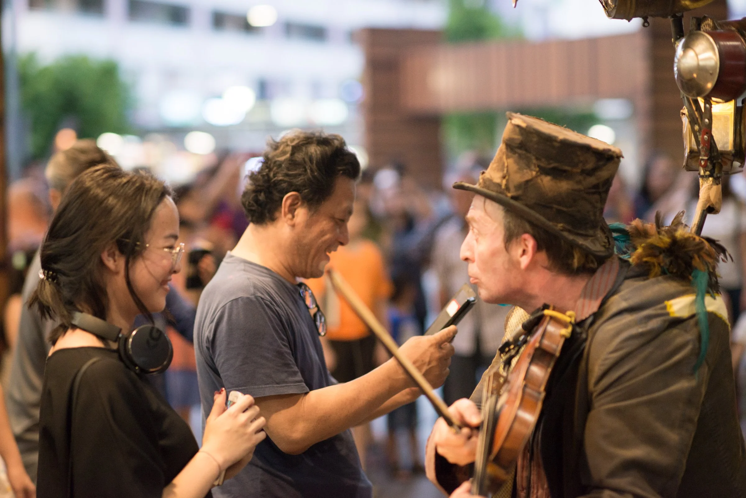 A woman with headphones around her neck and glasses laughing while talking to a man with a walking stick, another man wearing a tall, worn-out brown hat and playing a guitar, in a crowd at an outdoor event at dusk.