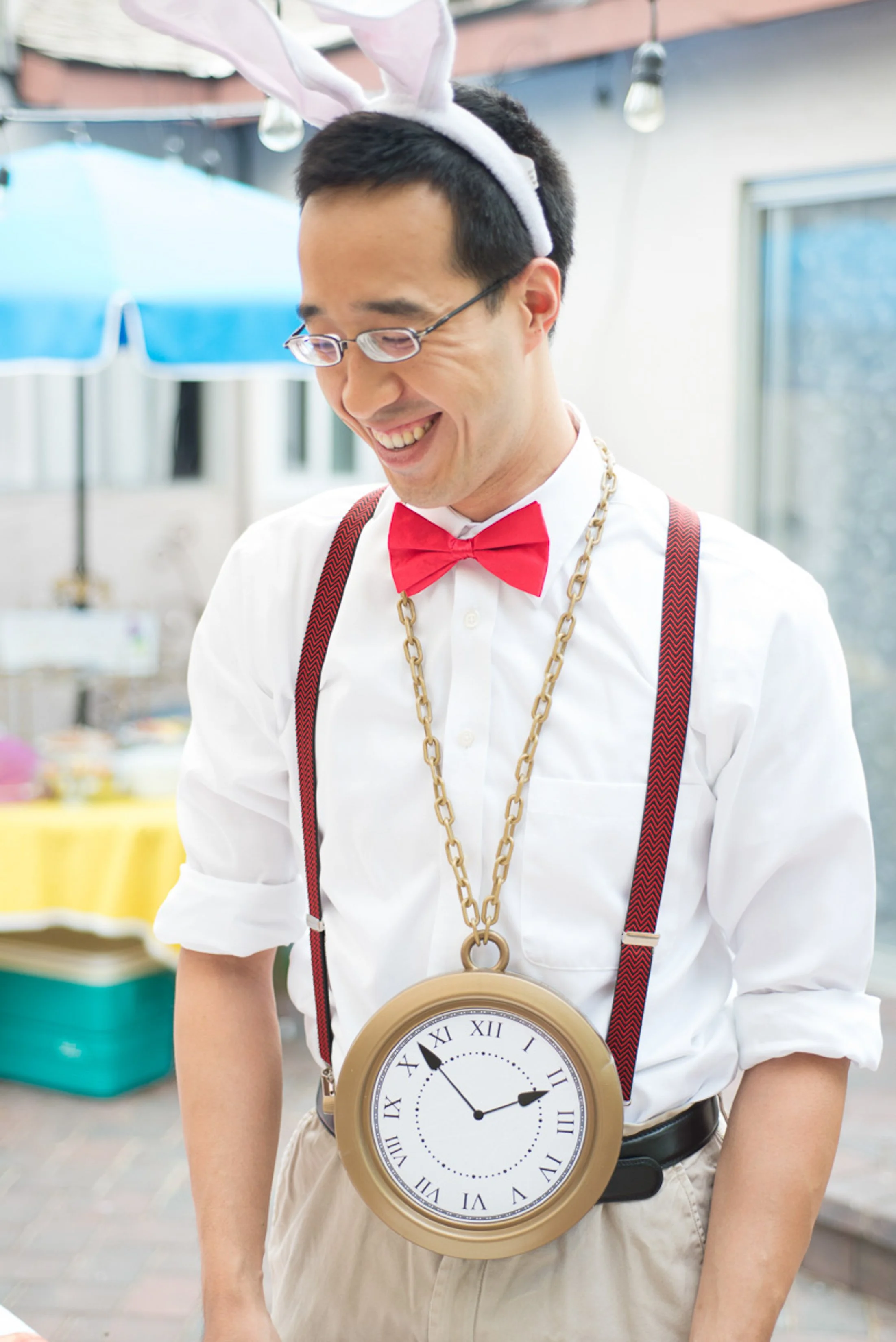 A man smiling wearing bunny ears, a red bow tie, suspenders, and a large clock necklace, standing outdoors with a blurred background.