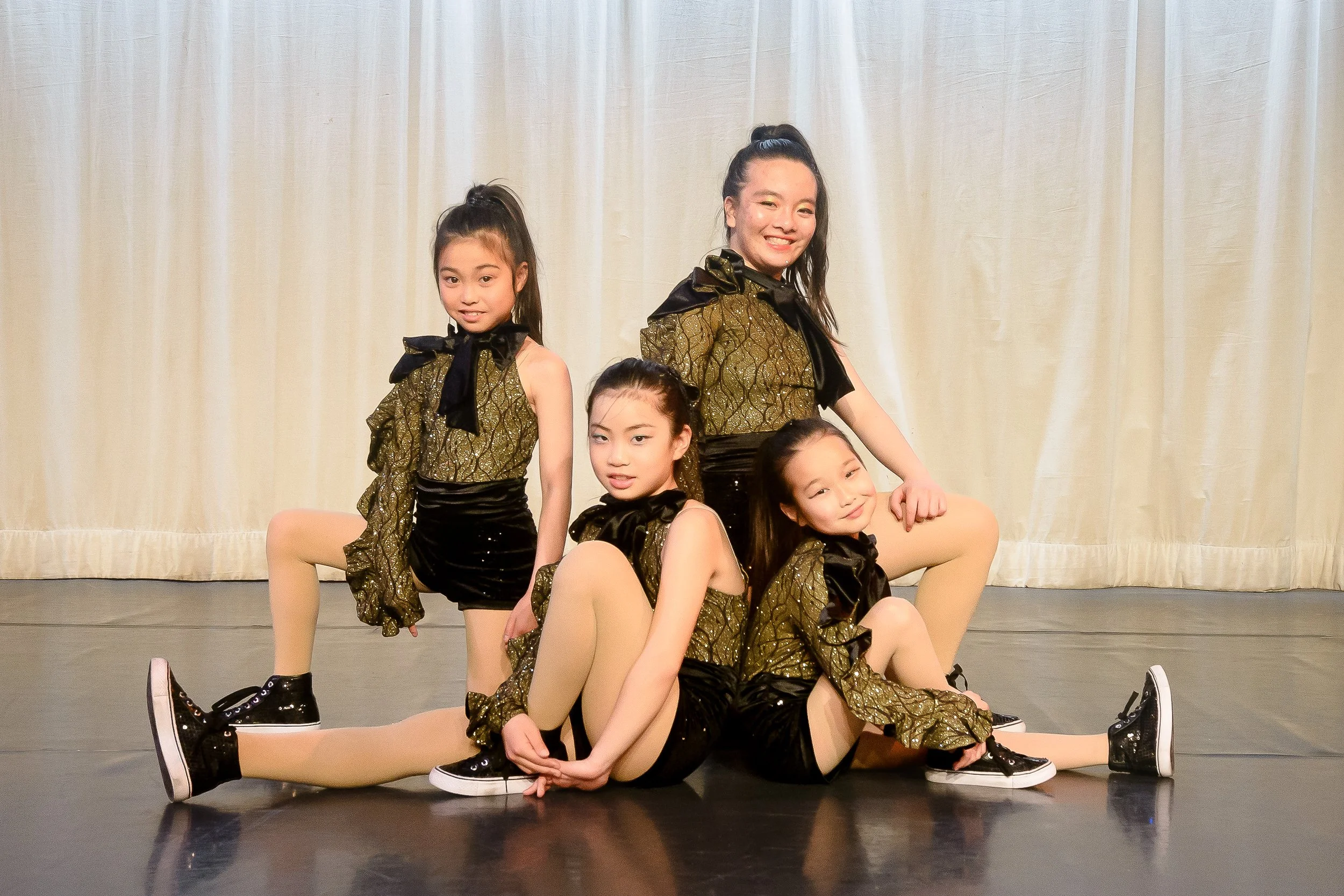 School photography. Five young girls dressed in matching gold and black dance costumes posing on stage in front of a beige curtain.
