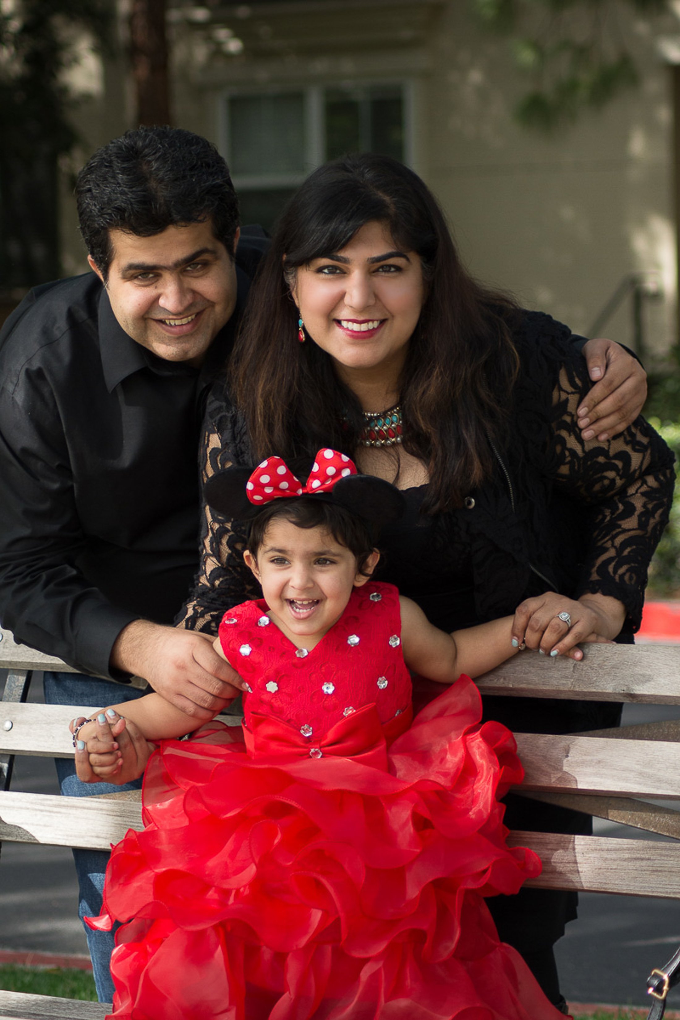 A happy family of three, a man, a woman, and a little girl, sitting on a park bench outdoors, smiling together. The girl is wearing a red dress with a large bow, and Minnie Mouse ears with a red polka dot bow.