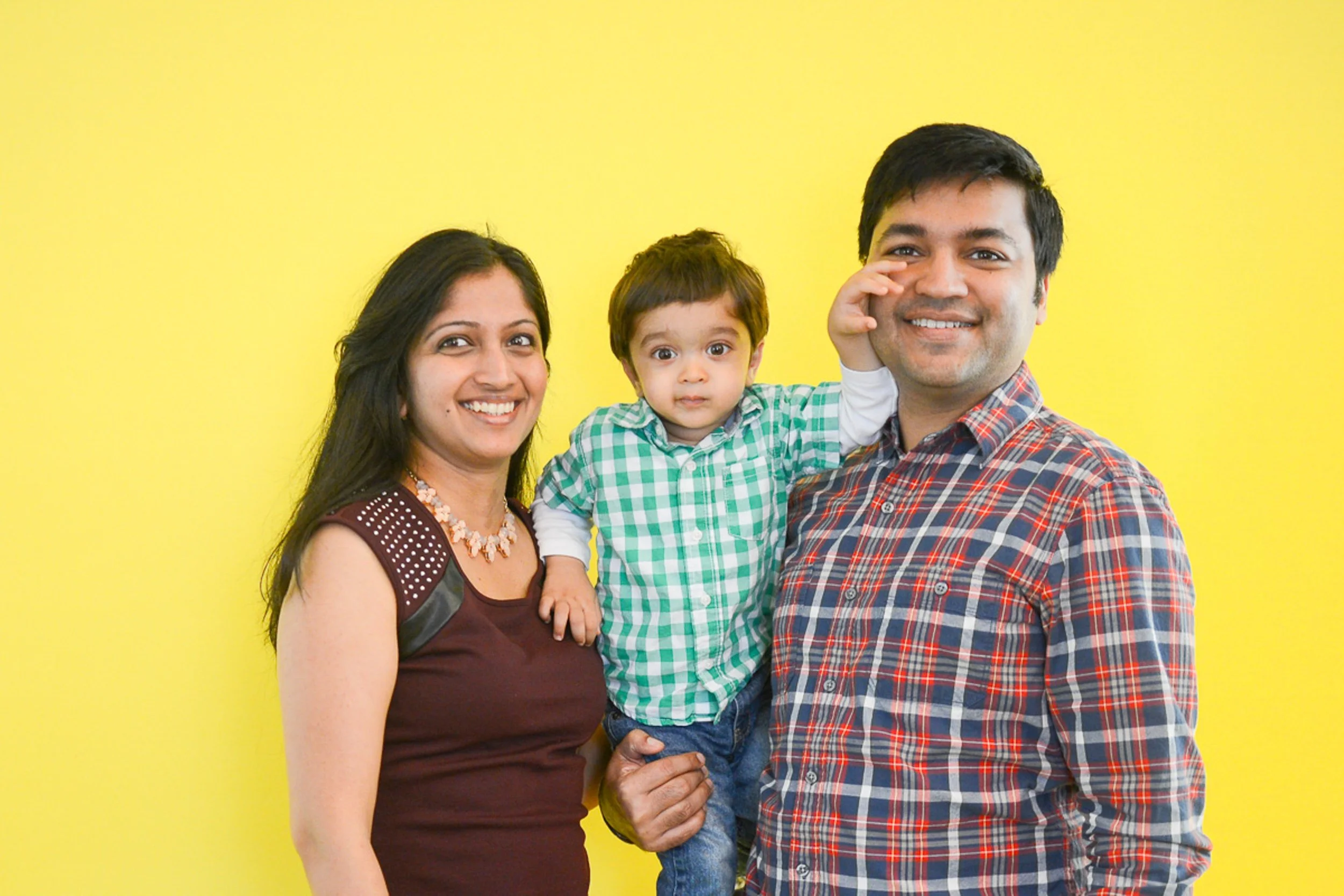 Happy South Asian family of three posing together against a yellow background, with a woman on the left, a man on the right, and a young boy in the middle.