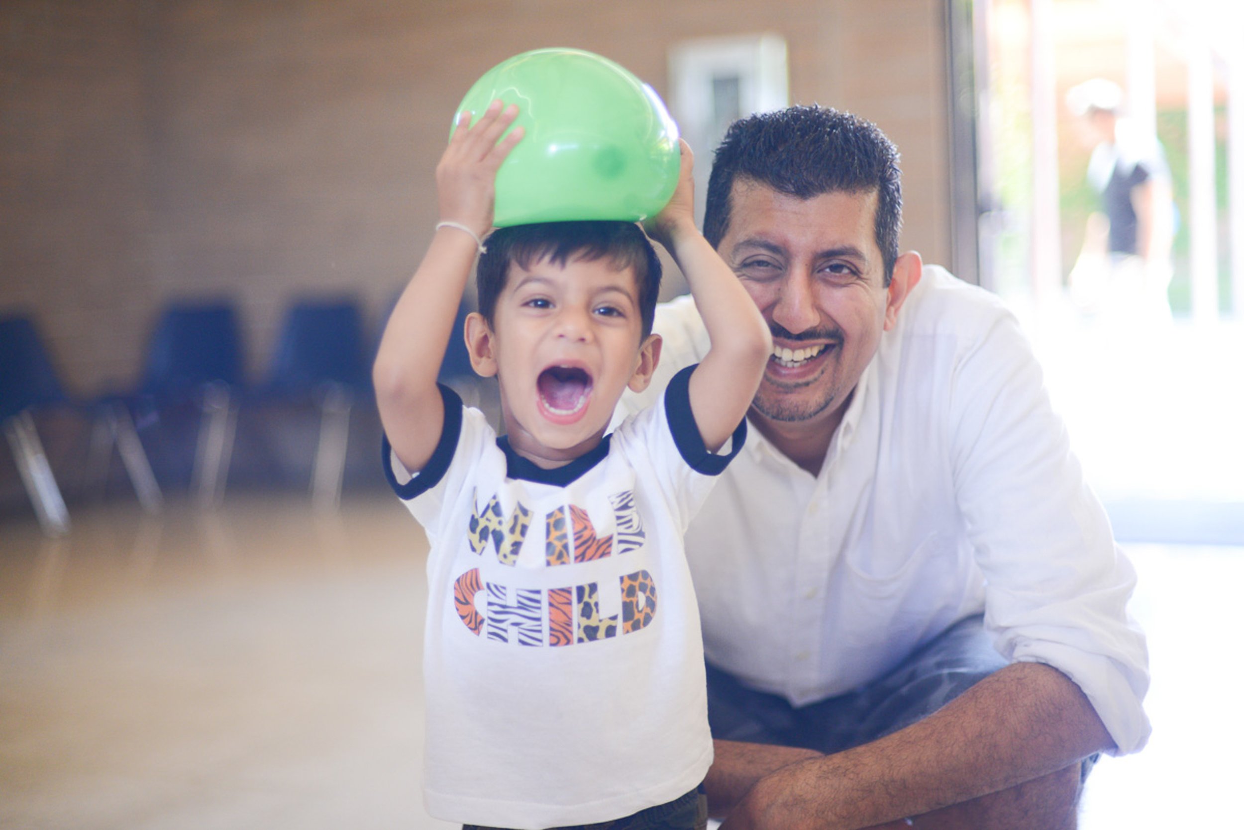 Event photography. A joyful young boy holding a green balloon on his head, standing next to a smiling man indoors near a window.