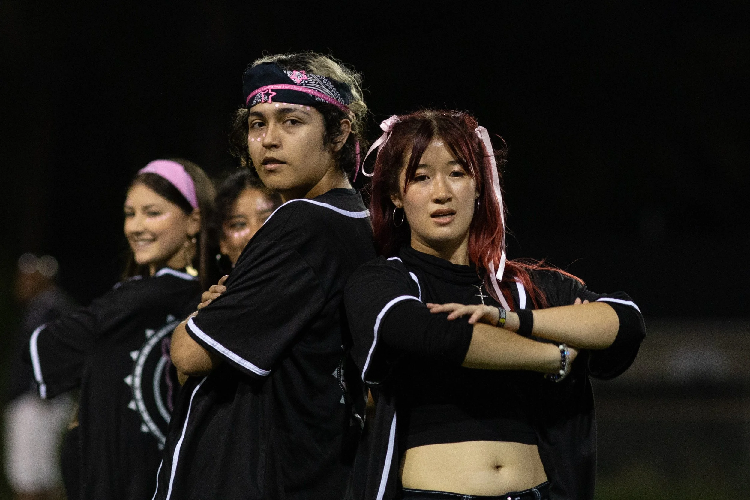 Four young women stand back-to-back at night, wearing black sports jerseys with white trim, posing with serious expressions and arms crossed or resting on their hips, outdoors under dark sky.