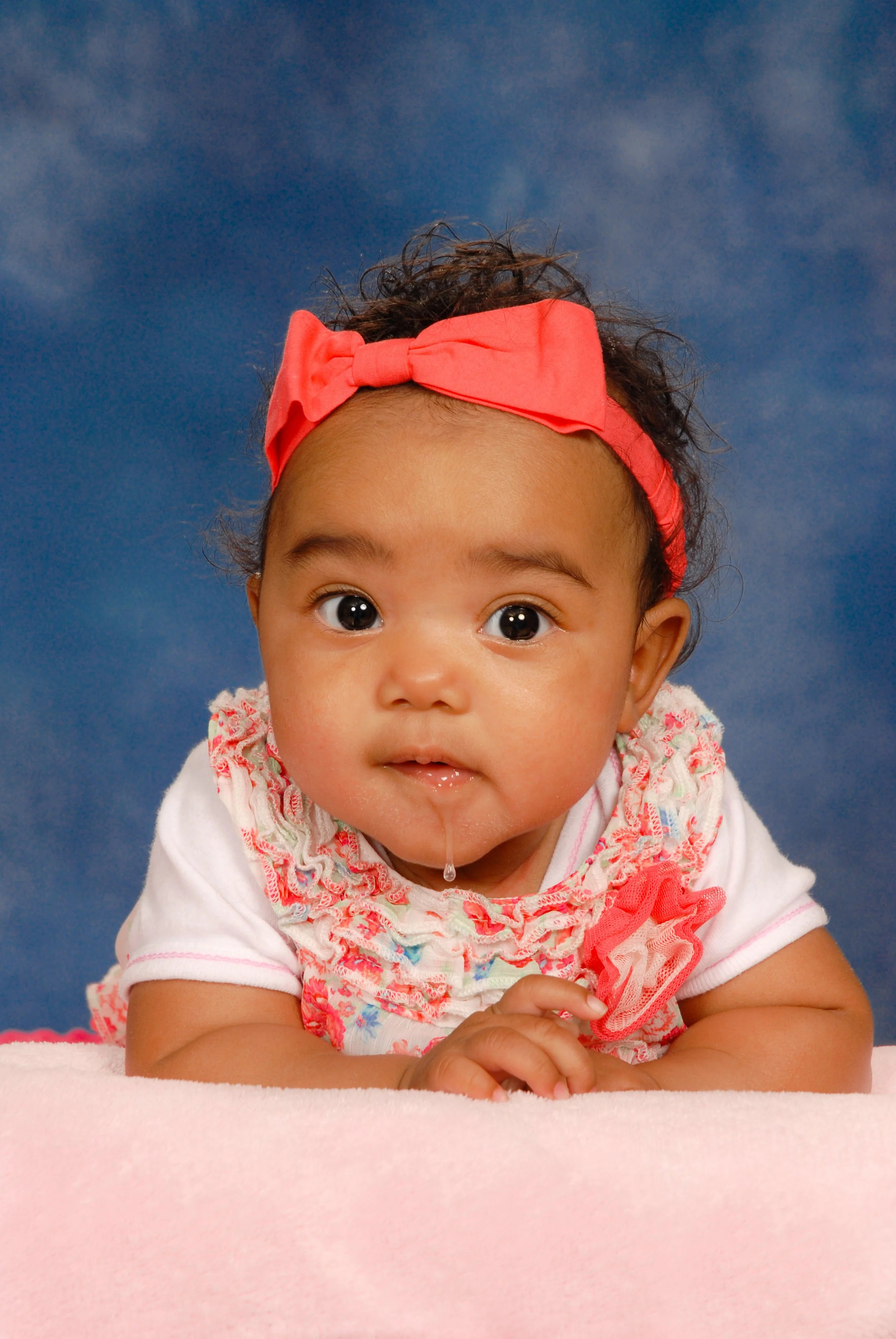 A baby girl with a red bow headband, lying on her stomach on a pink surface, against a blue background, with big eyes and a slight drool on her chin.