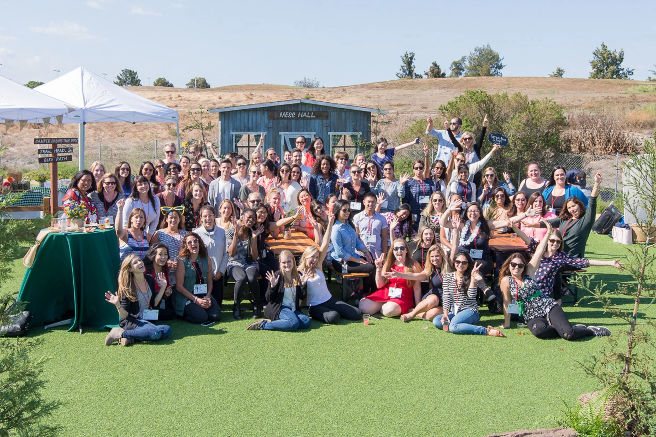 Large group of people outdoors on a sunny day, sitting and standing on green grass with a rustic blue building labeled 'Mess Hall' in the background. Some individuals are waving and smiling at the camera, with a tent and a sign indicating directions 