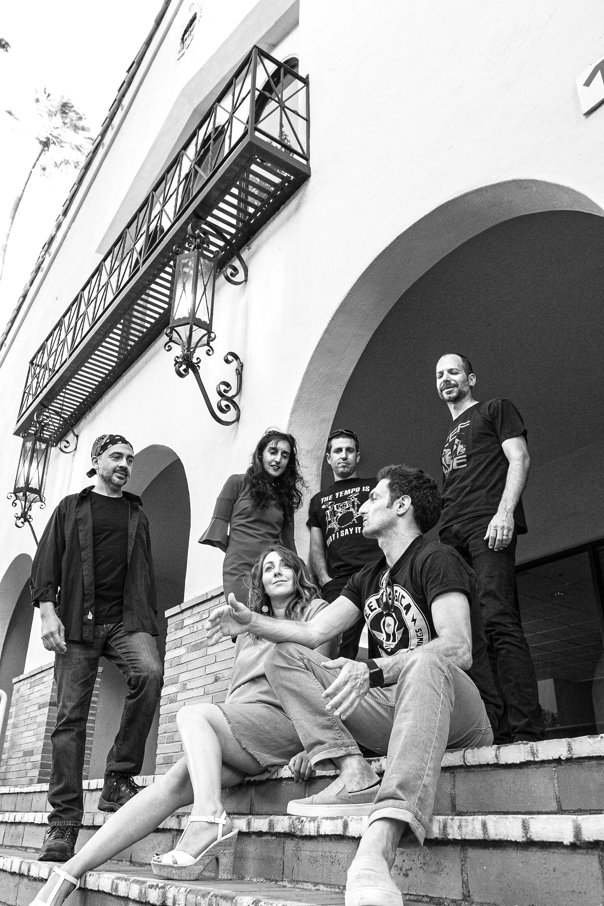 Group of six people, three men and three women, gathered on stairs outside a building with arches. One woman is seated on the stairs, gesturing as she talks. Others stand nearby, engaging and looking at her. The setting features a building with a bal