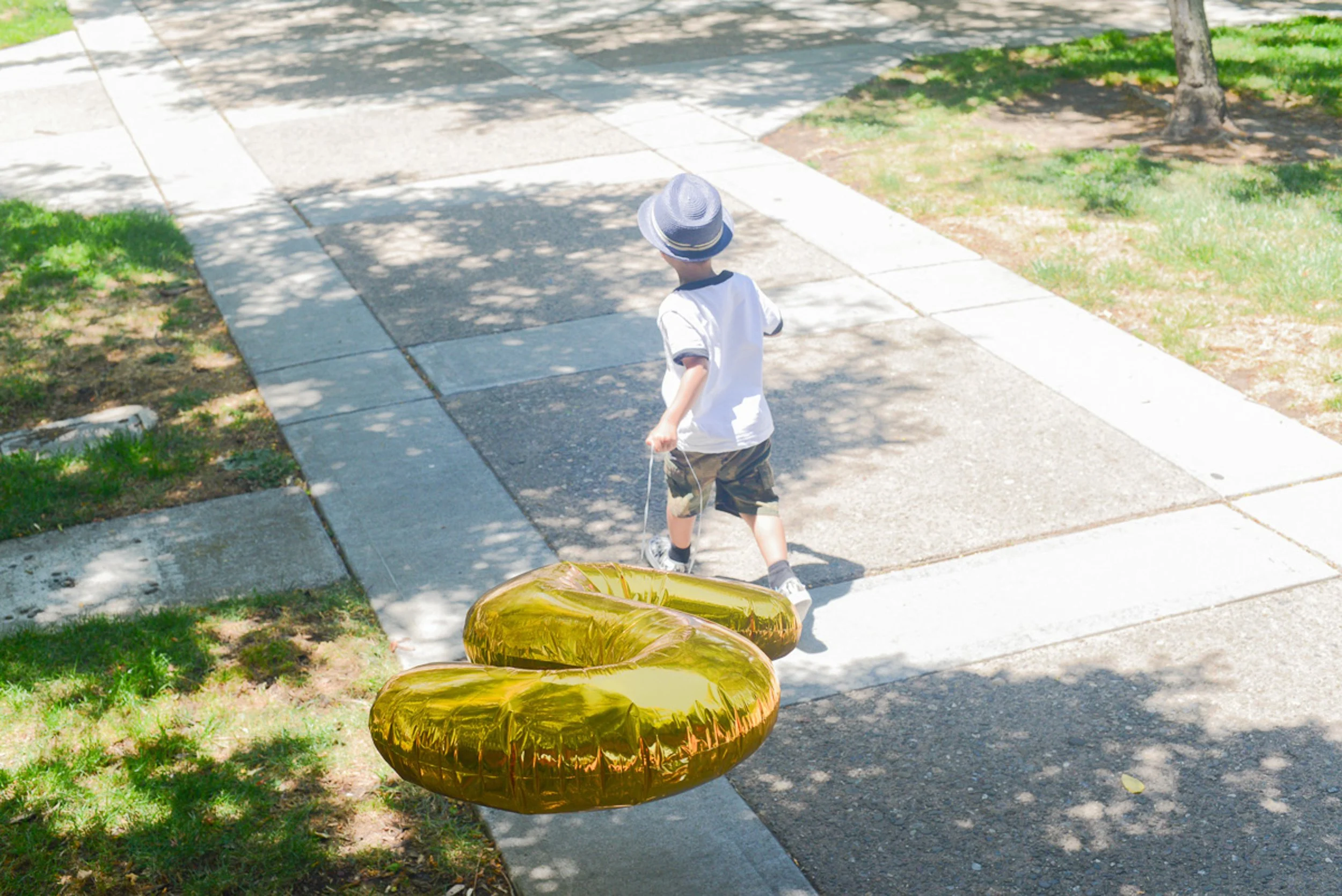 A young boy wearing a white shirt, camouflage shorts, and a wide-brimmed hat walks on a sidewalk while pulling a gold balloon shaped like the number 2.