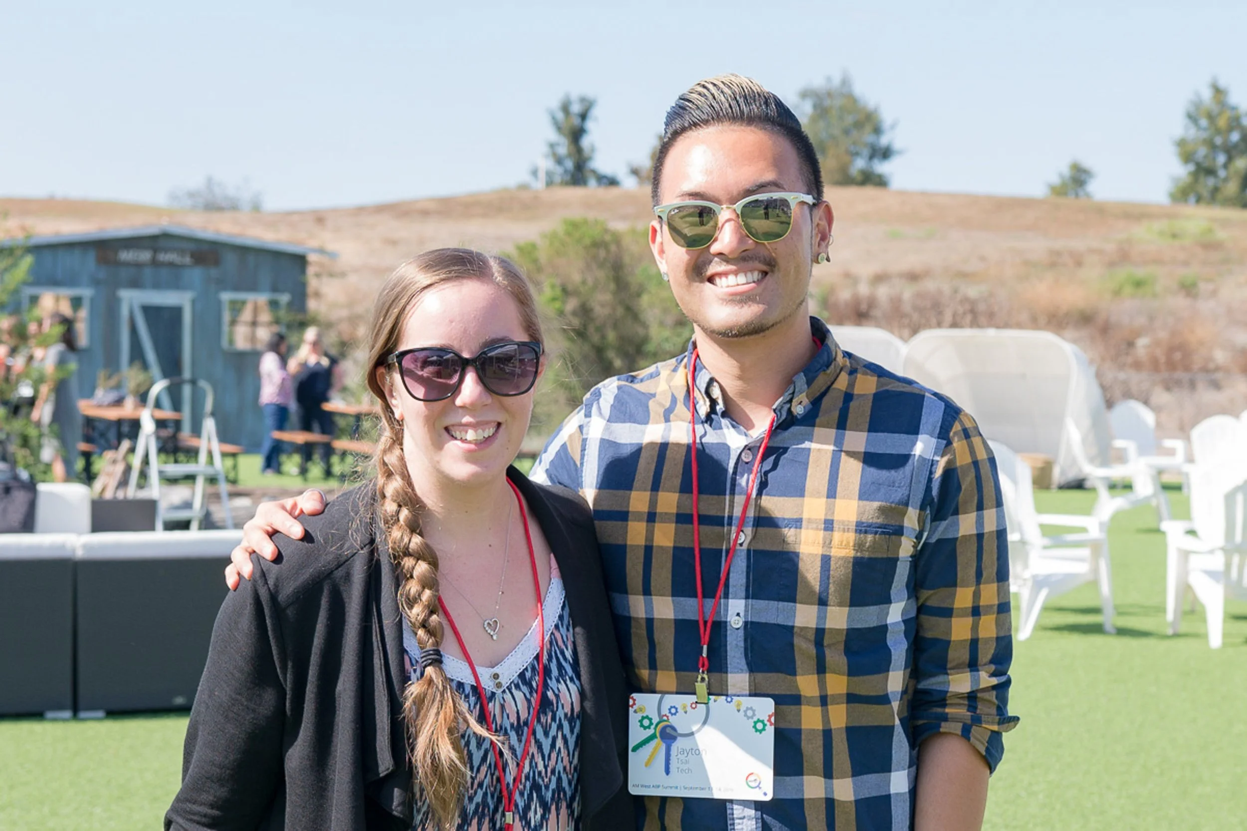 A smiling man and woman wearing sunglasses and name tags, standing outdoors on a sunny day with green grass and a hill in the background.