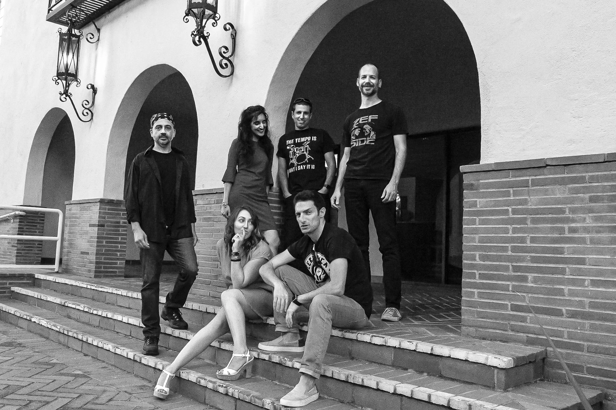 Group of seven people posing on steps outside a building with arched doorways, some standing and some sitting, in black and white photograph.