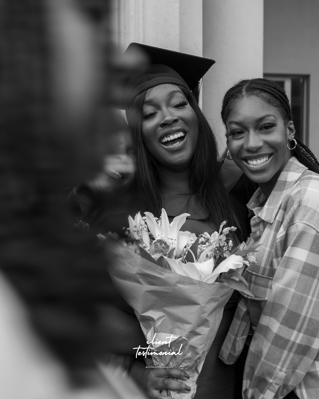 Black and white photo of a young woman in a graduation cap smiling with two women, one holding a bouquet of flowers, celebrating graduation.