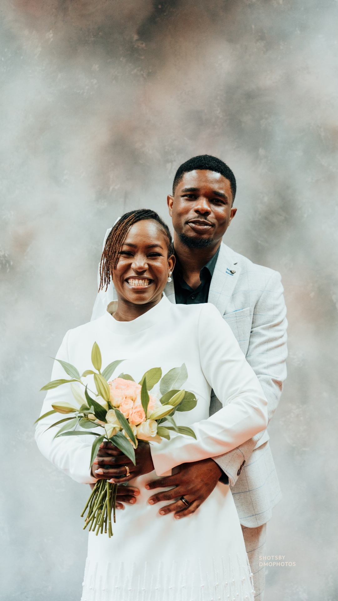 A smiling woman wearing a white dress holding a bouquet of flowers, standing in front of a man in a light gray suit with a black shirt, both standing against a gray textured background.