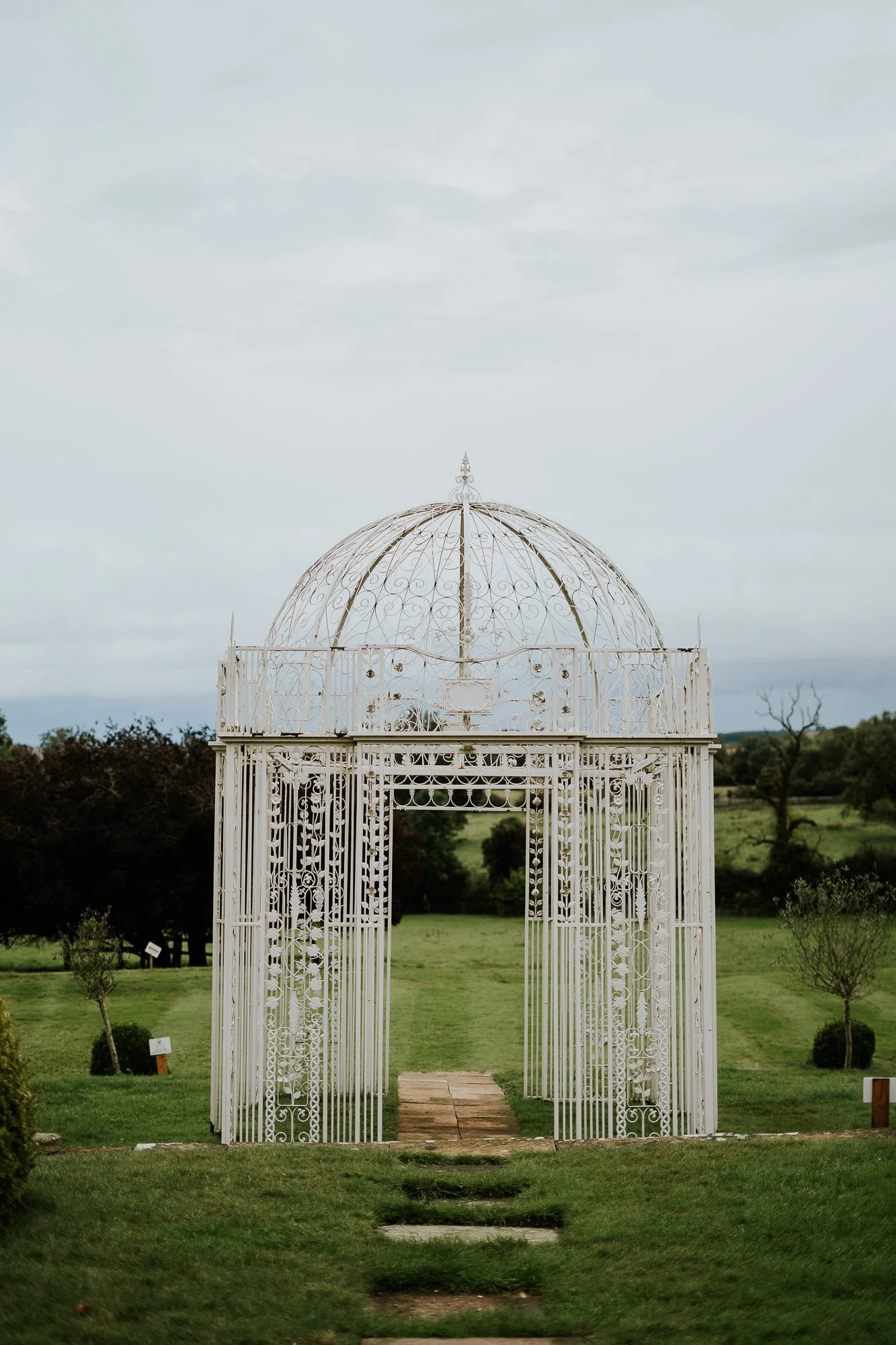 A white ornate iron gazebo on a green lawn with trees and cloudy sky in the background.