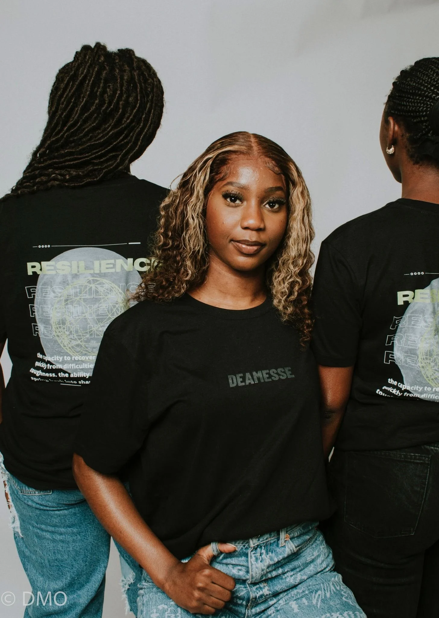 Three women wearing black shirts with the word 'Resilience' on the back, standing close together. The woman in the middle faces forward, with blonde curly hair and a confident expression. The other two women face away, showing their hairstyles with p