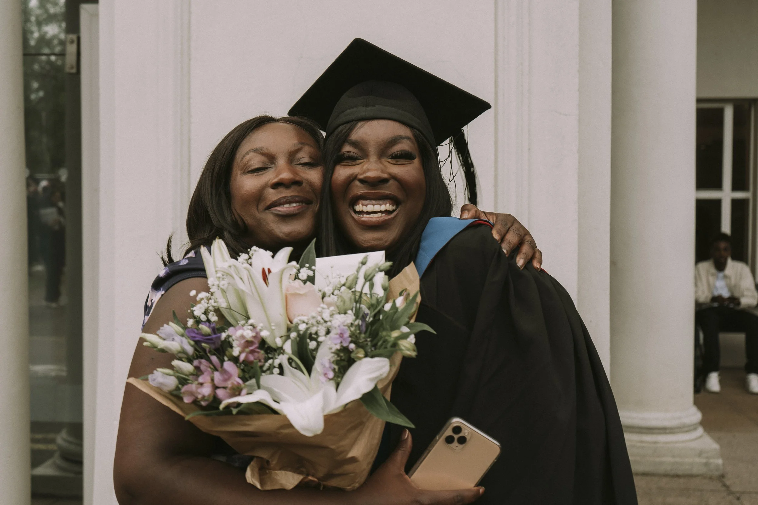 A woman in graduation cap and gown is hugging another woman, holding a bouquet of flowers and a smartphone, as they smile and celebrate.