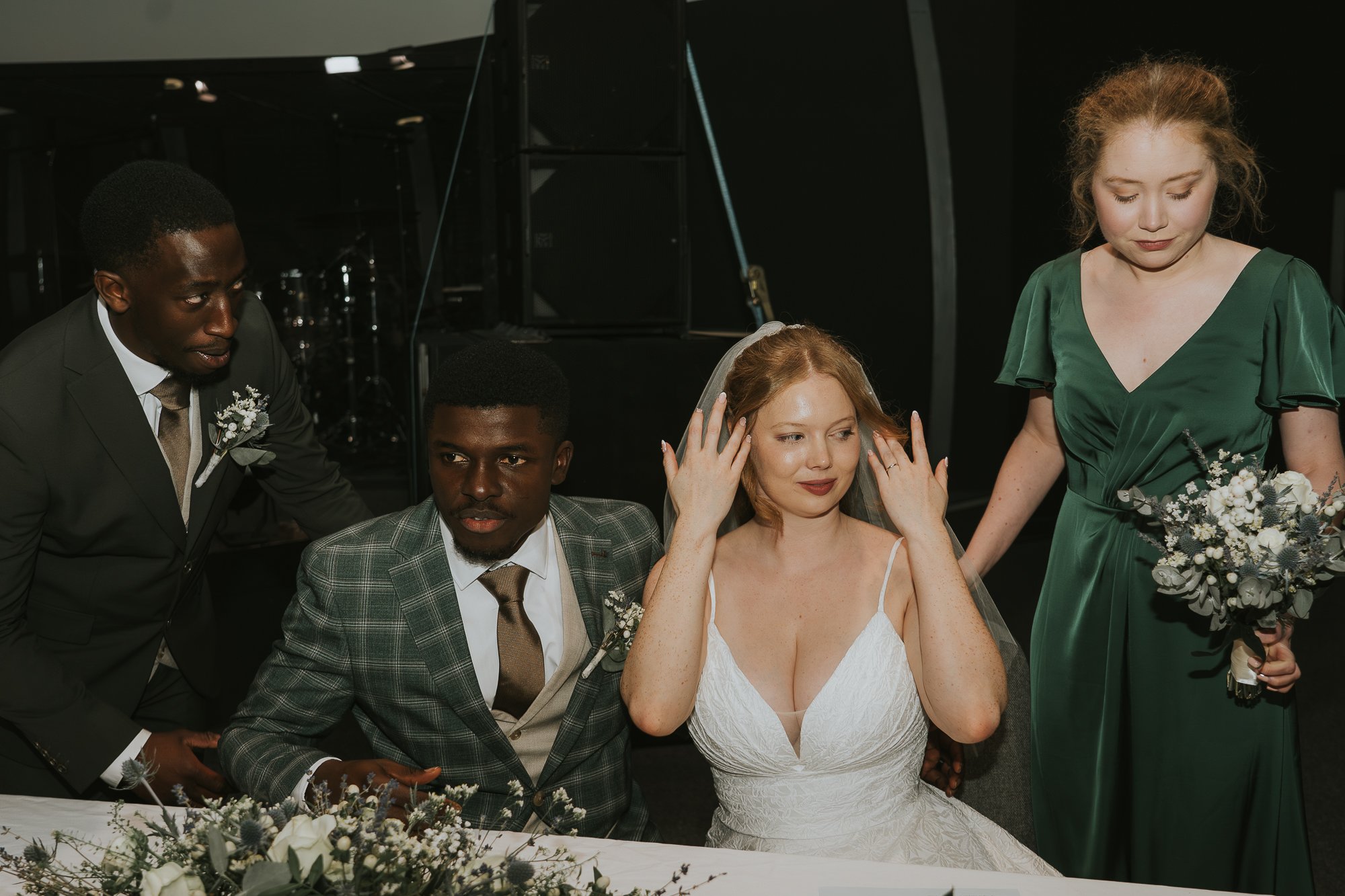 A bride seated at a wedding table, touching her hair, with a man in a plaid suit and a woman in a green dress standing beside her, and two men in suits sitting nearby.