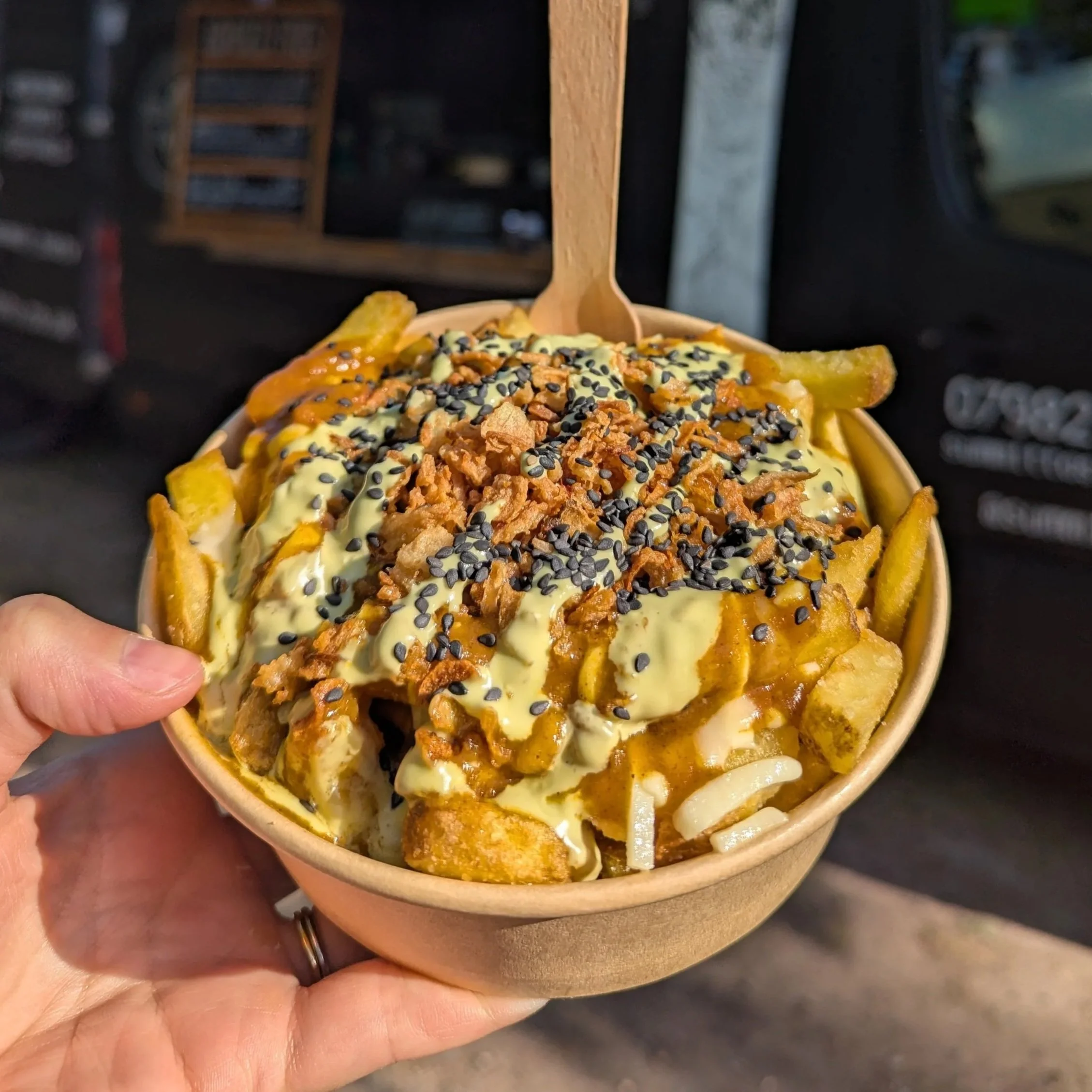 Skin on fries topped with a rich homemade katsu curry, plant-based chicken chunks, tangy pickled Carrots, crunchy edamame beans, crispy panko breadcrumbs and a sprinkle of black sesame seeds held up against the Summit Tasty food truck