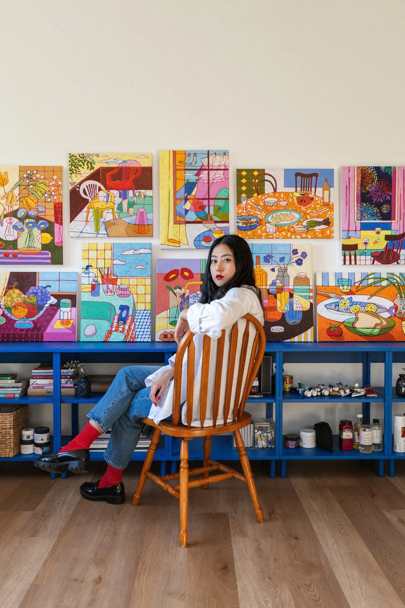 A young woman with dark hair, wearing a white shirt, jeans, red socks, and black shoes, sitting on a wooden chair in front of a blue shelf filled with art supplies and books, with colorful paintings on the wall behind her.