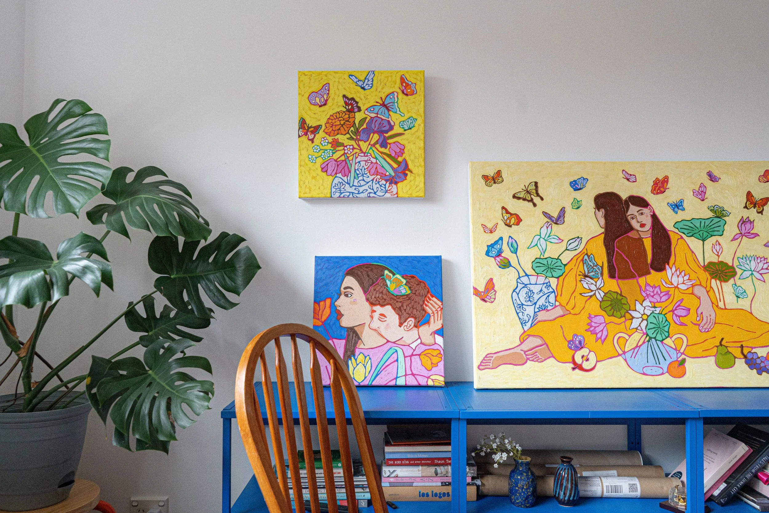 Interior of an art display with three colorful paintings on a white wall, large green monstera plant in a gray pot to the left, a blue table with books, vases, and small decorative items, and wooden chair partially in view.