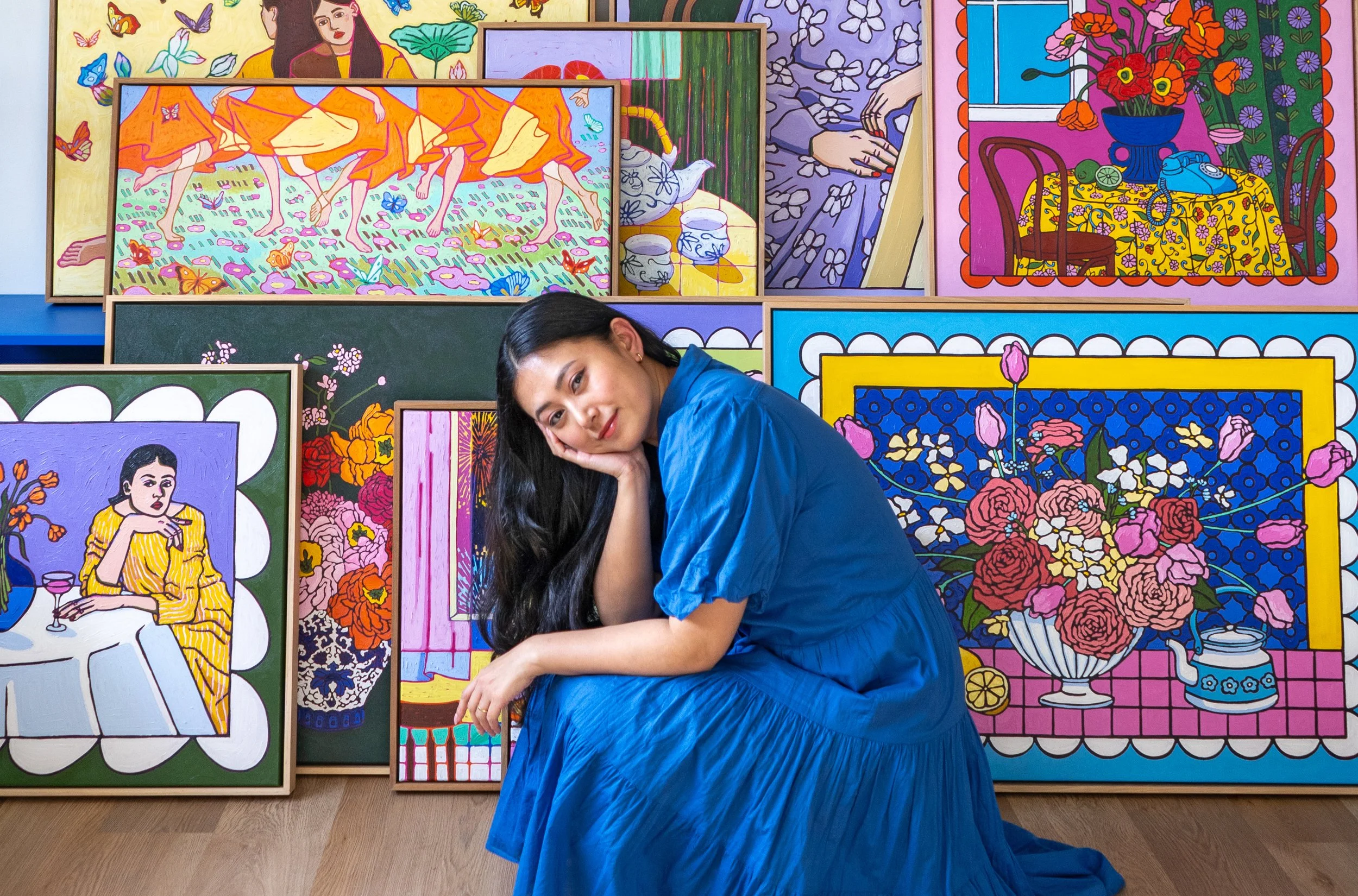 A woman in a blue dress sitting on the floor with her head resting on her hand, surrounded by colorful framed paintings of floral and portrait scenes.
