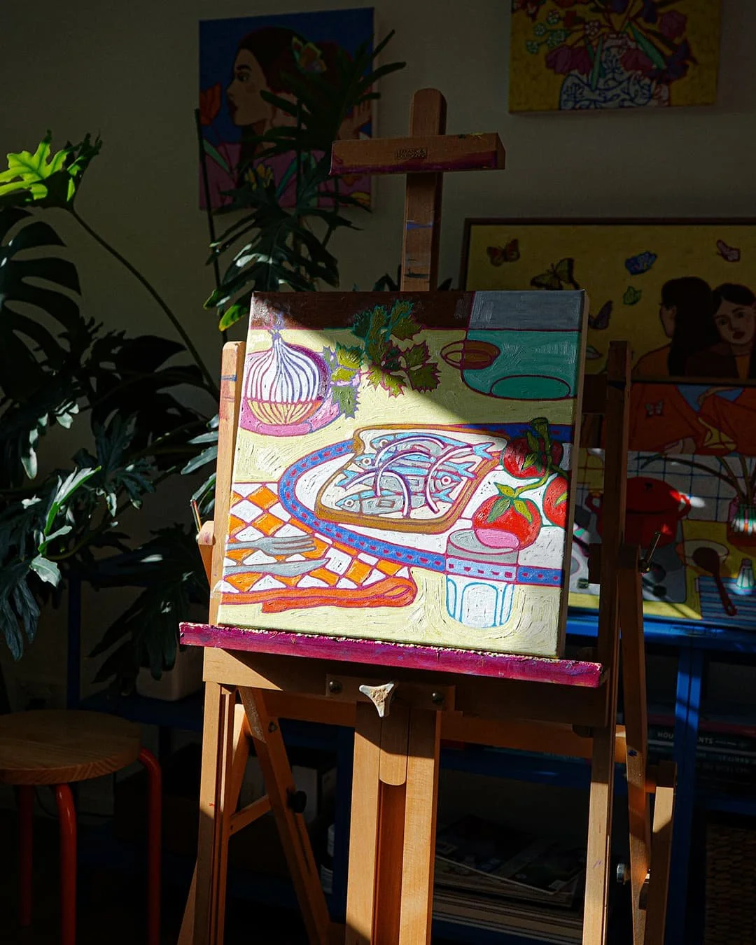 Colorful still life painting of vegetables and table setting on an easel in a room with plants and other artwork.