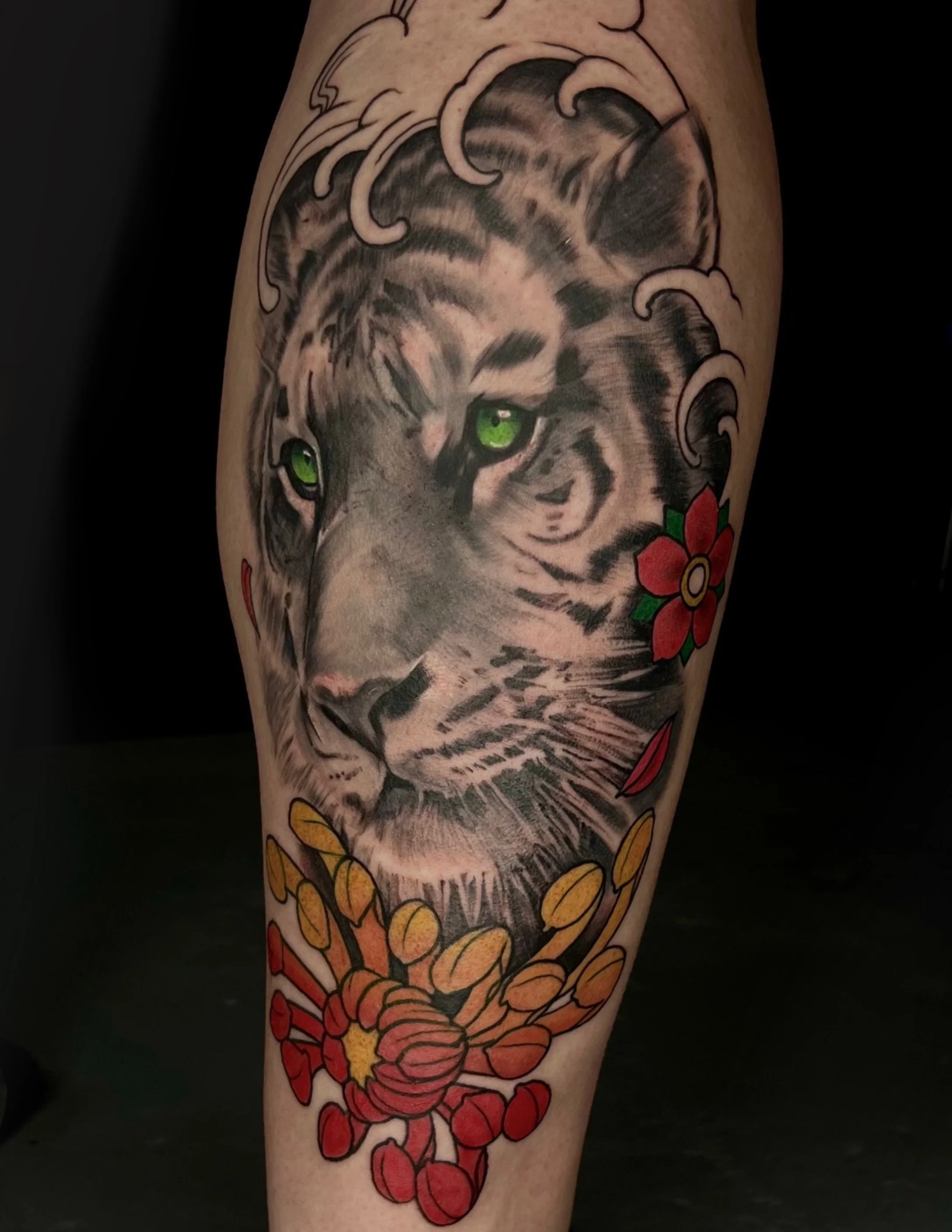 Tattoo of a tiger's face with vivid green eyes, surrounded by red, orange, and yellow flowers and leaves.