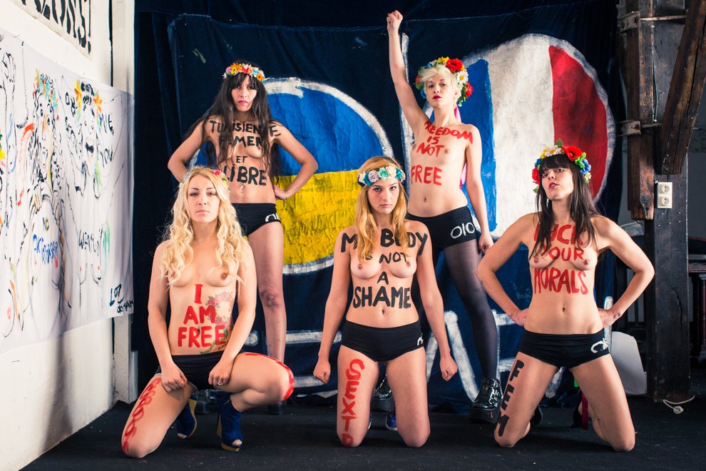 Cinq jeunes femmes debout ou agenouillées, portant des couronnes de fleurs, affichent des messages écrits sur leur corps, dans un décor artistique avec un fond coloré et une toile illustrée.