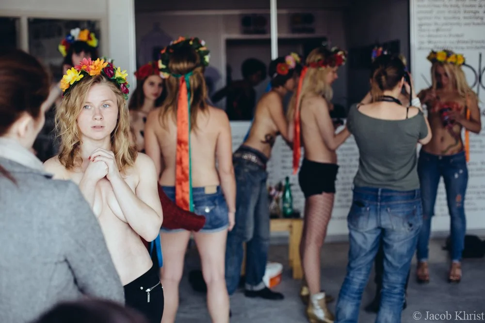 Jeune femme nue portant une couronne de fleurs, pensant ou anxieuse, face à une autre femme habillée, dans un environnement de coulisses ou de répétition, avec plusieurs femmes en arrière-plan portant également des couronnes de fleurs et des vêtement