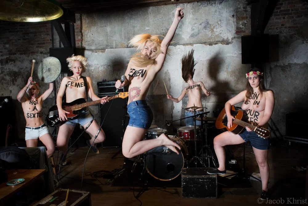 Un groupe de jeunes femmes nues avec des inscriptions écrites sur leur corps, jouant de la musique dans un lieu industriel.