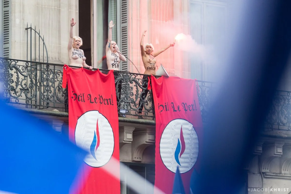 Manifestantes topless en una protesta, sosteniendo banderas rojas con un símbolo y el texto 'Oui Le Pen', en un edificio con balcones de hierro forjado.