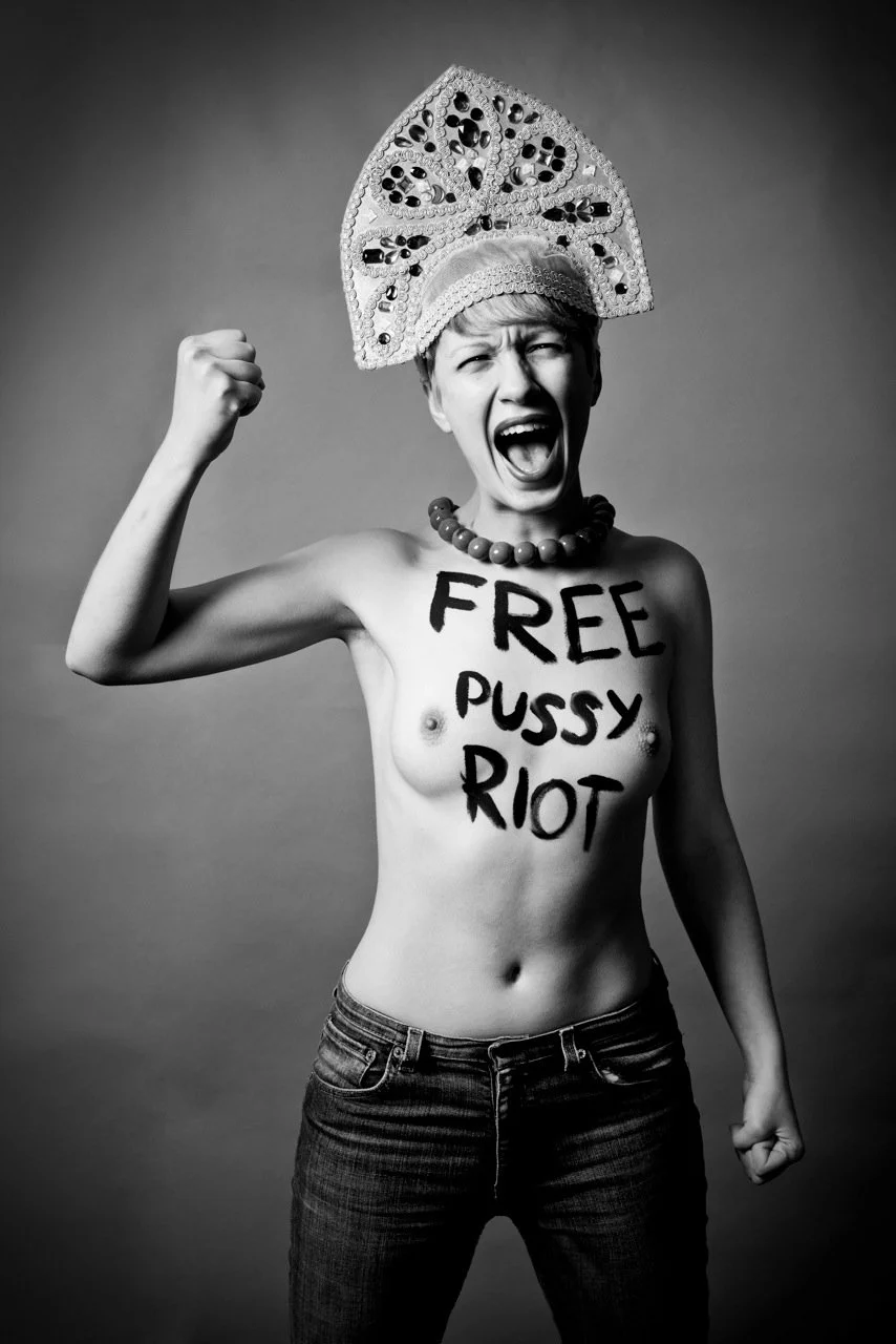 Une femme topless portant un grand couvre-chef décoré, faisant un geste de défi, avec le texte "FREE PUSSY RIOT" peint sur son corps.