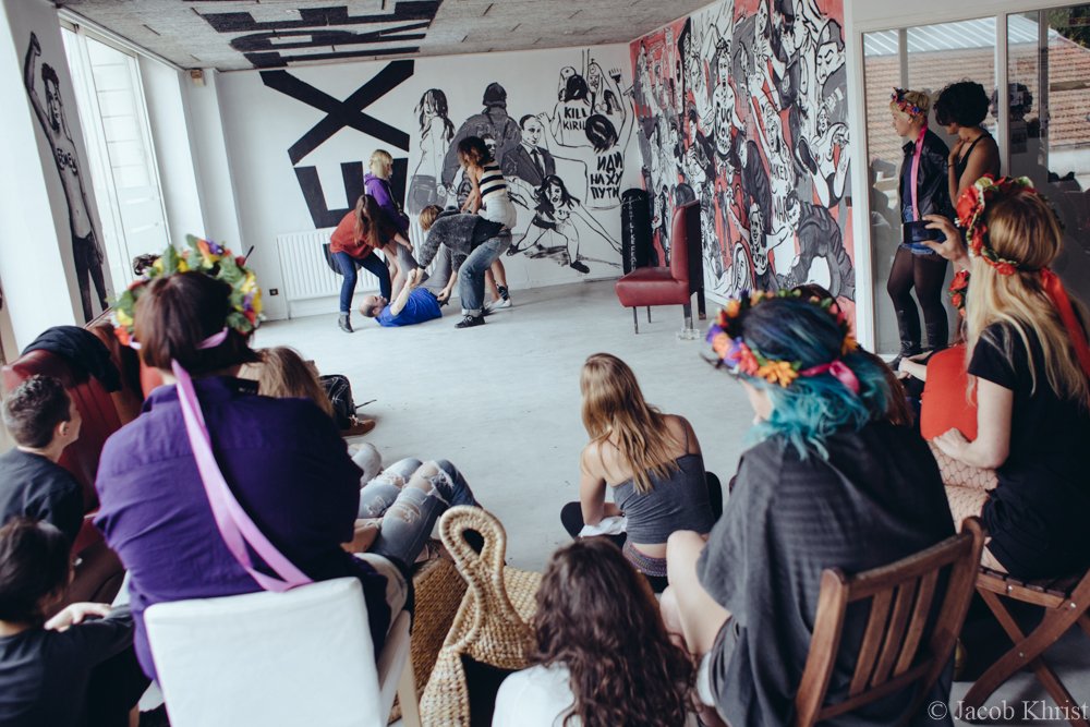 Une scène de spectacle ou de mise en scène de violence dans une salle avec mur de graffitis, des spectateurs regardant, certains portant des couronnes de fleurs.